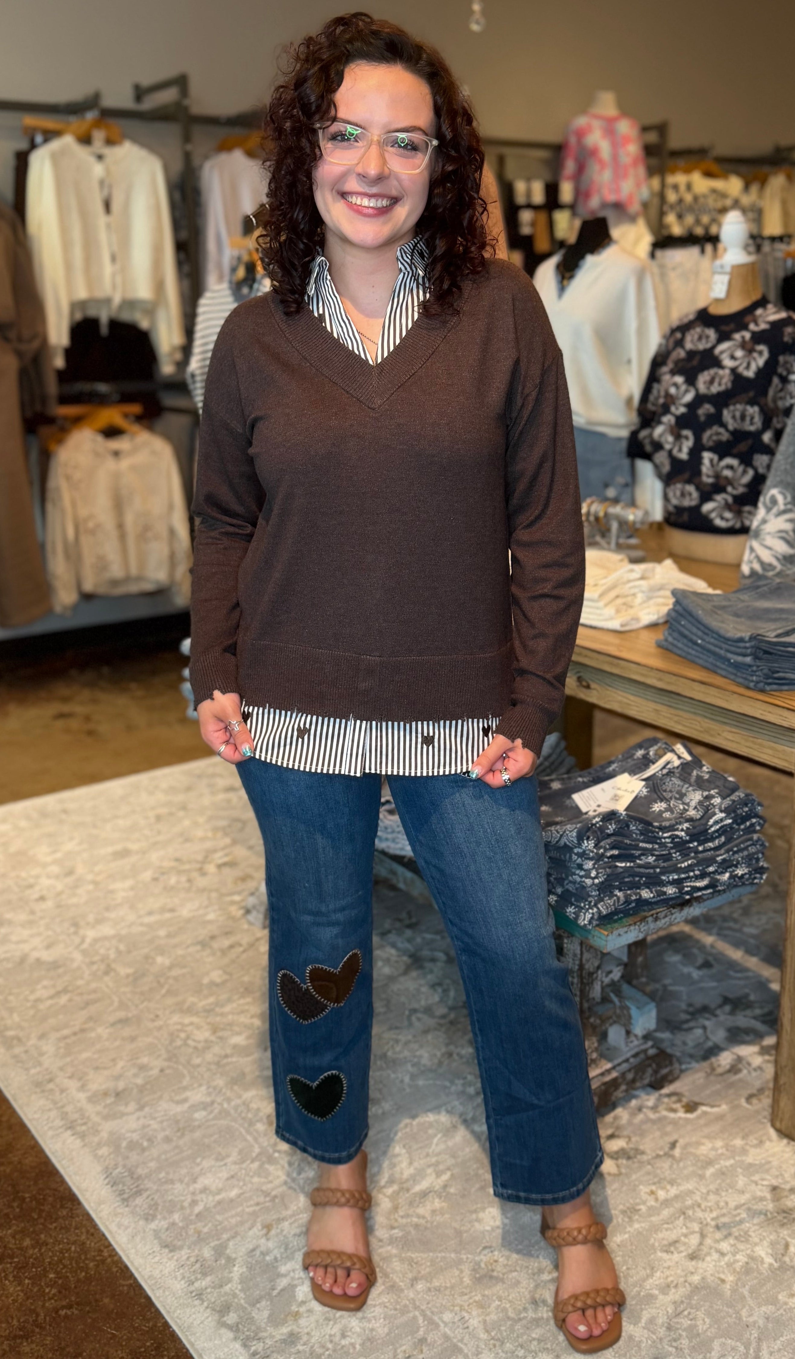 A woman stands in a clothing boutique wearing a brown v-neck sweater, a striped shirt, and blue jeans with heart patches. Clothing racks, a table with folded garments, and a patterned rug are visible in the background.