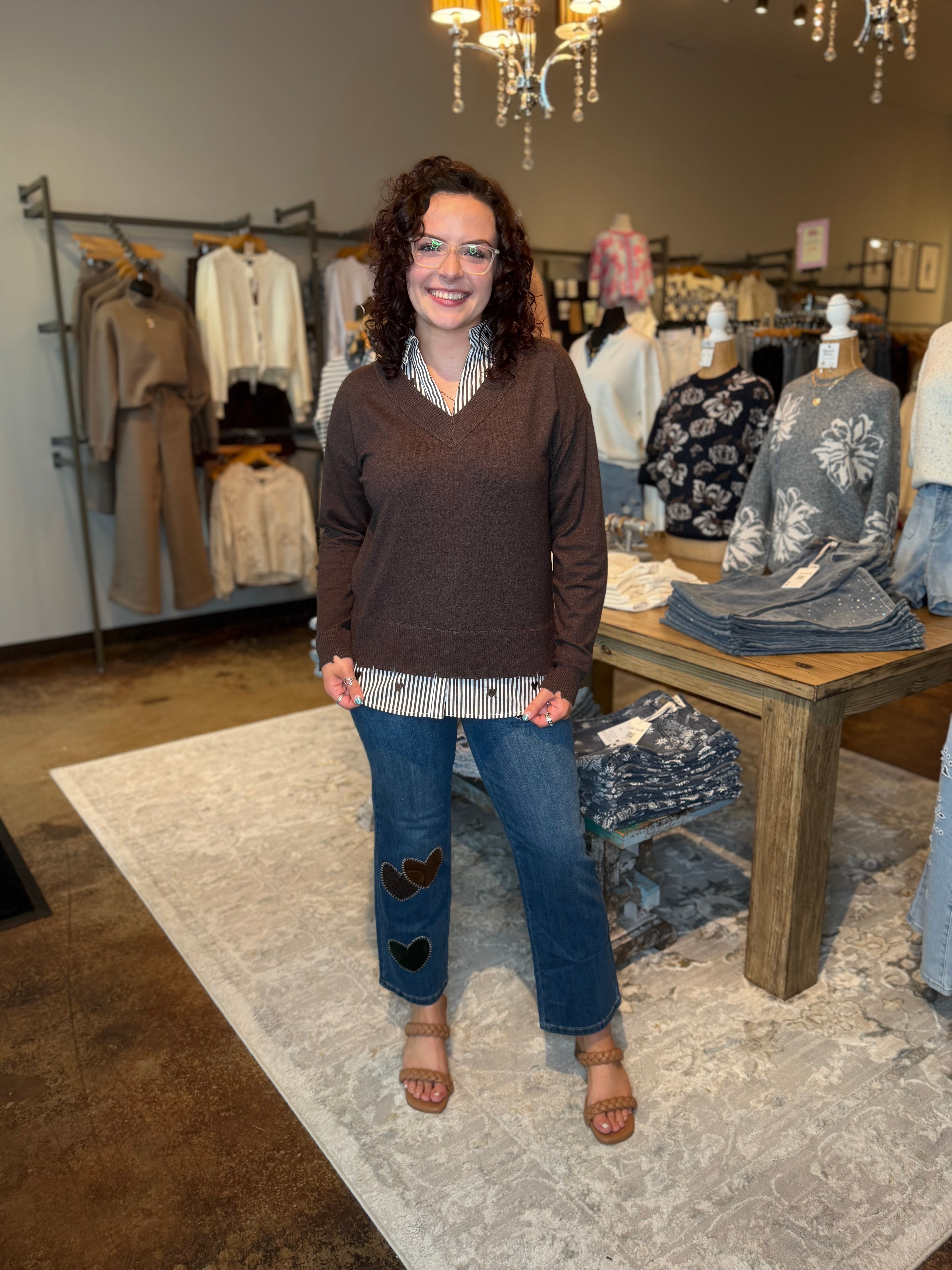 A woman stands in a clothing boutique wearing a brown v-neck sweater, a striped shirt, and blue jeans with heart patches. Clothing racks, a table with folded garments, and a patterned rug are visible in the background.