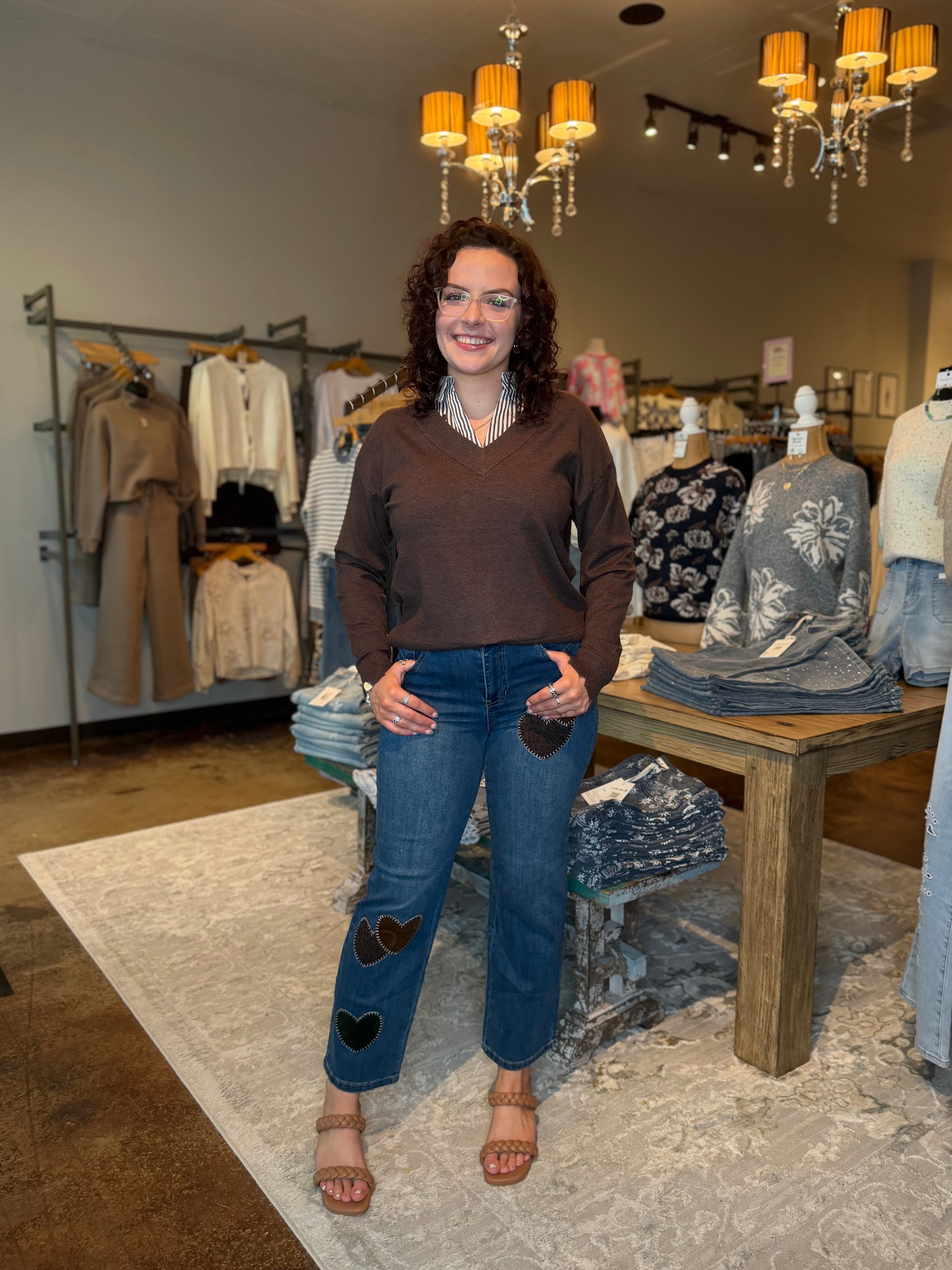 A woman stands in a boutique wearing a brown long-sleeve top and blue jeans with heart-shaped patch details on the knees. The store has clothing racks, a wooden table with folded garments, and hanging orange lamps.