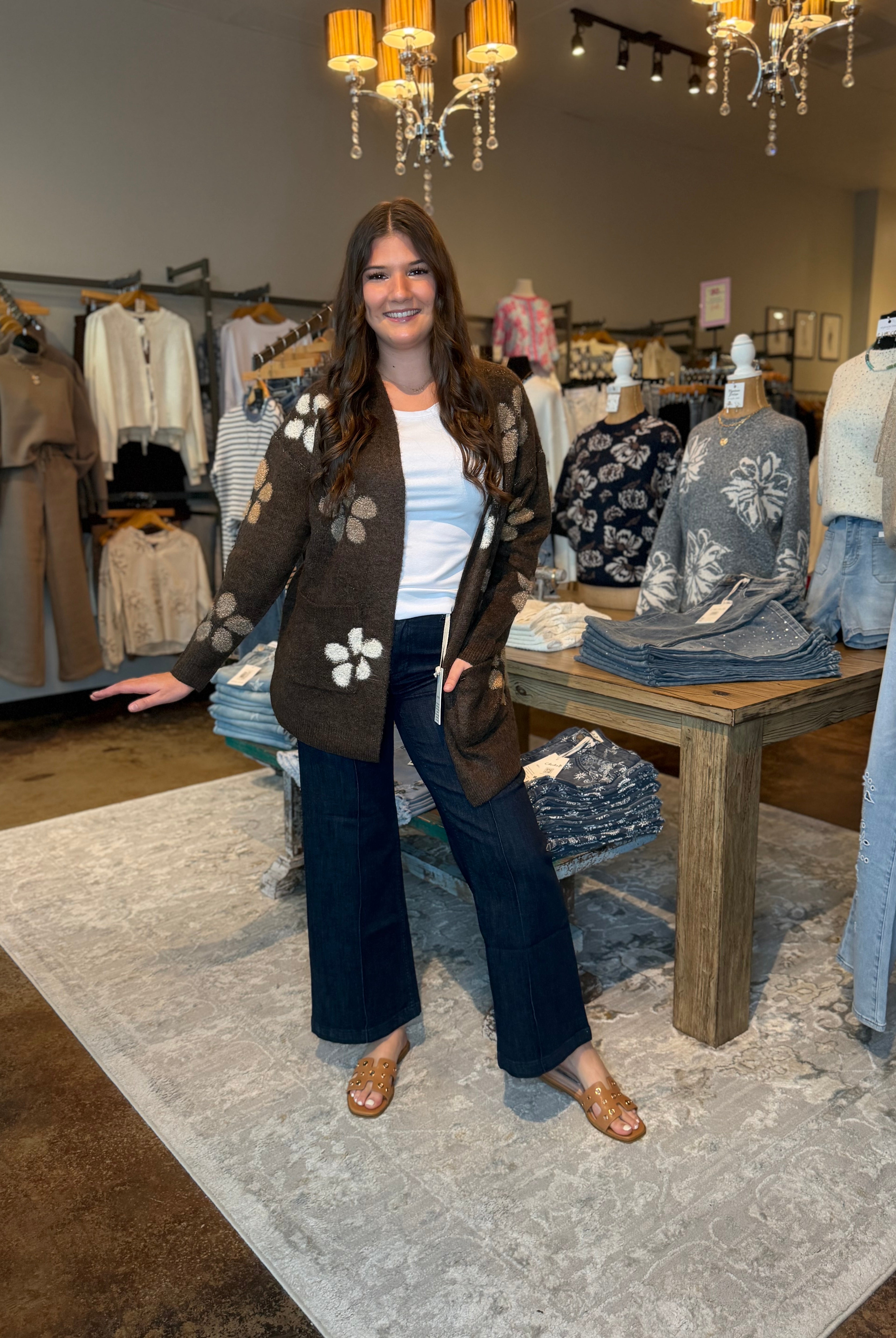A woman stands in a boutique wearing a brown knit cardigan with white floral motifs, a white top, dark wide-leg pants, and brown flat shoes. The background shows clothing racks, mannequins, a wooden table with folded garments, and hanging lights.