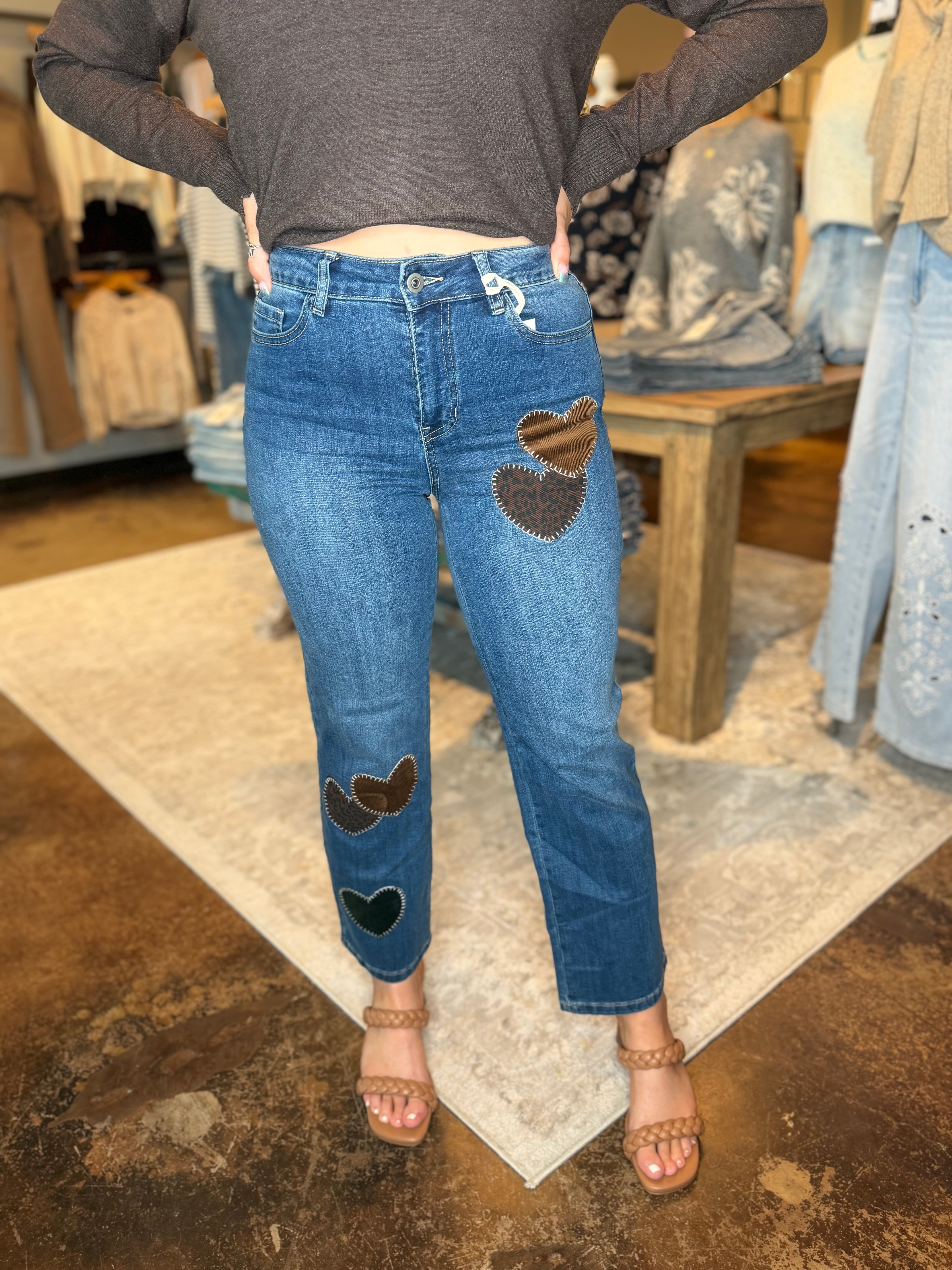 A person wearing blue jeans with brown heart patches on the front and lower legs, paired with a dark gray long-sleeve crop top and tan heeled sandals, standing in a clothing store.