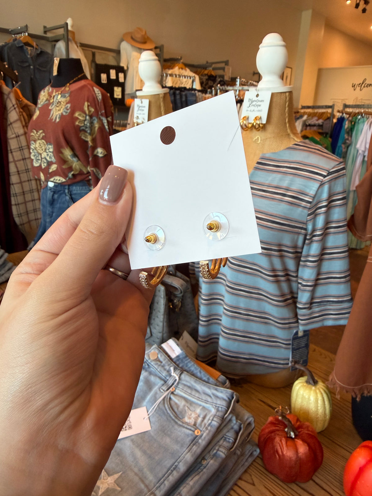 A hand holds a white display card featuring two small gold and white stud earrings with clear round backs, set against a busy boutique background with mannequins and clothing racks.