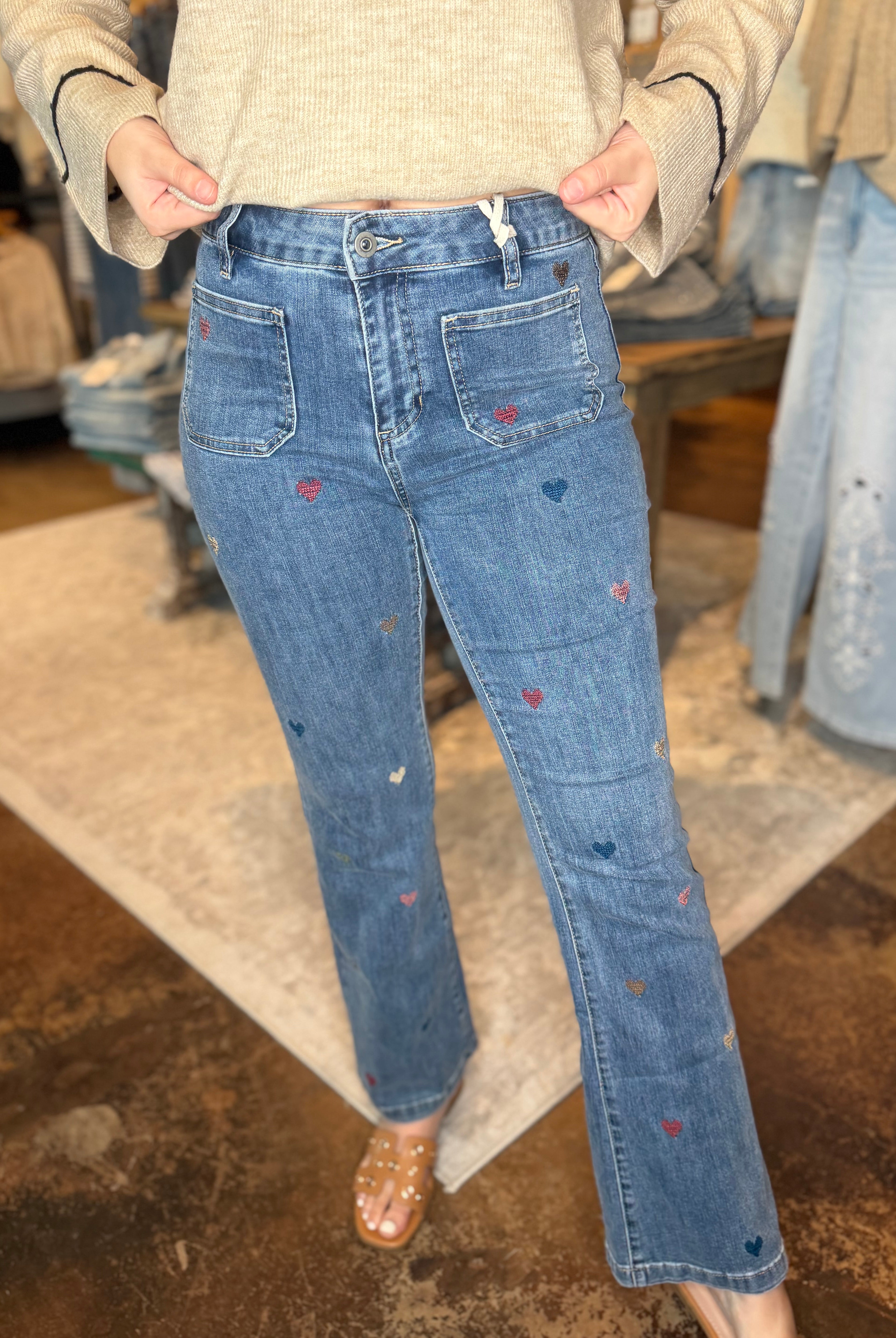 A person wearing blue denim jeans with small embroidered heart details in red, blue, and white, paired with a beige knit sweater and clear flat sandals in a retail store setting.