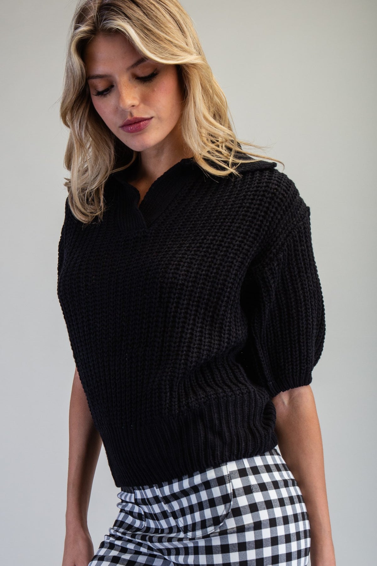 A woman wearing a black textured short-sleeve knit top and a black-and-white gingham skirt, photographed against a plain light gray studio background.
