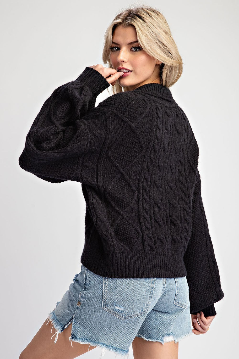 A woman wearing a black cable-knit sweater and light-wash denim shorts, photographed against a plain light gray studio background.