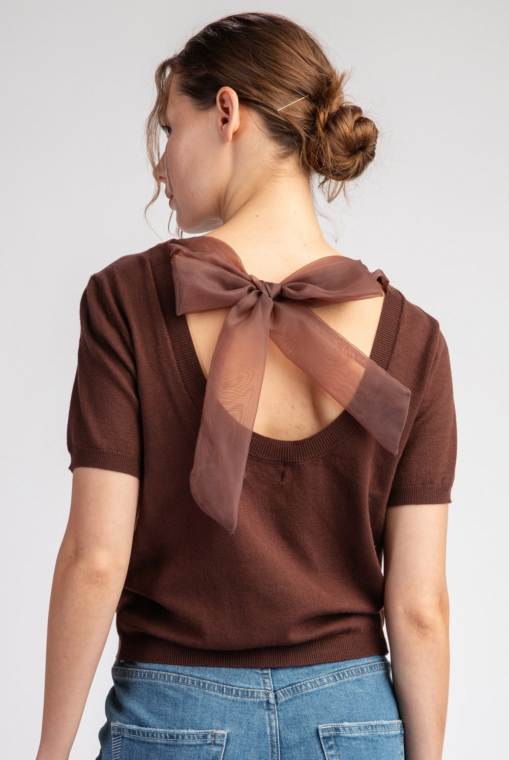 Woman wearing a brown blouse with a bow detail on the back against a plain background