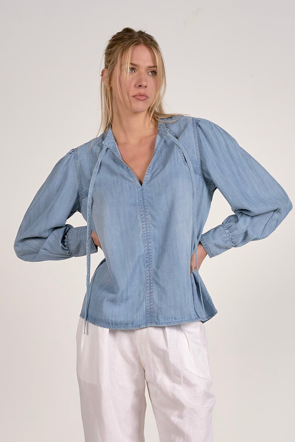 A woman modeling a light blue chambray long-sleeve blouse with a split V-neck and drawstring tie details, paired with white pants, against a plain light gray studio background.