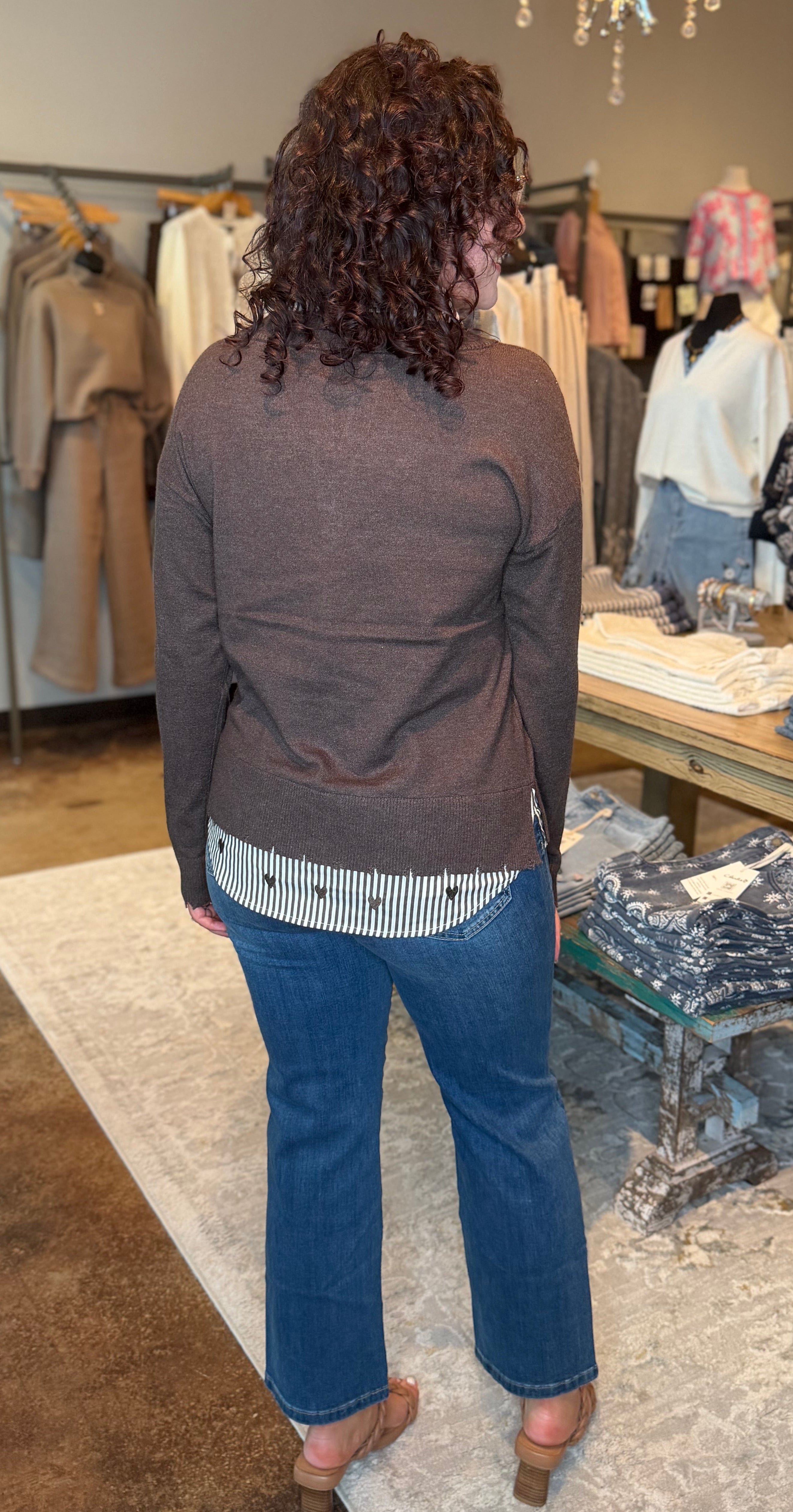 A woman viewed from behind wearing a brown knit cardigan, a striped shirt, and blue jeans inside a clothing boutique.