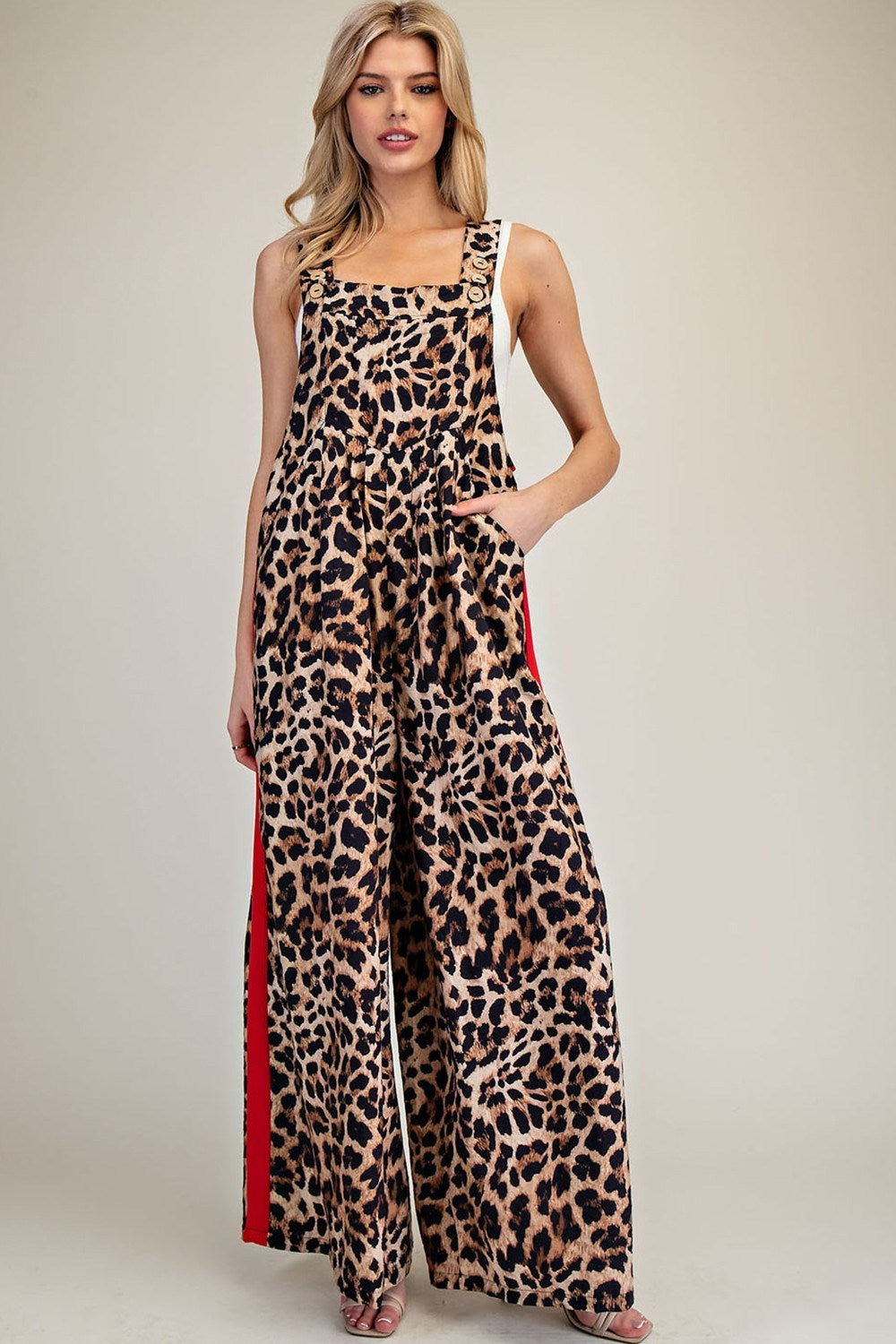 A woman modeling a leopard-print sleeveless jumpsuit with wide legs and red side panels, photographed against a plain light beige studio background.