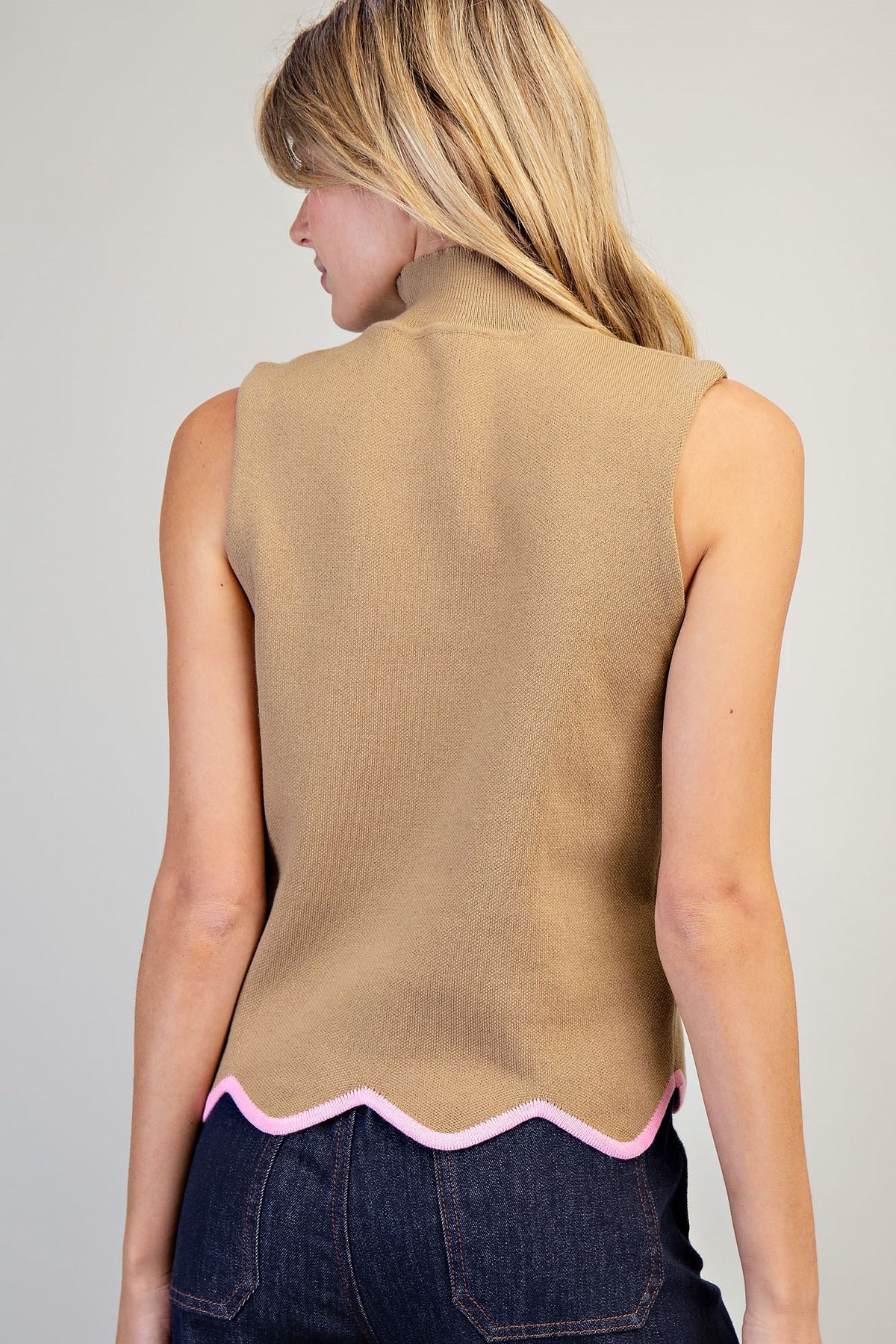 A woman shown from the back wearing a sleeveless camel-colored knit top with a pink scalloped hem, paired with dark blue jeans, against a light gray studio background.