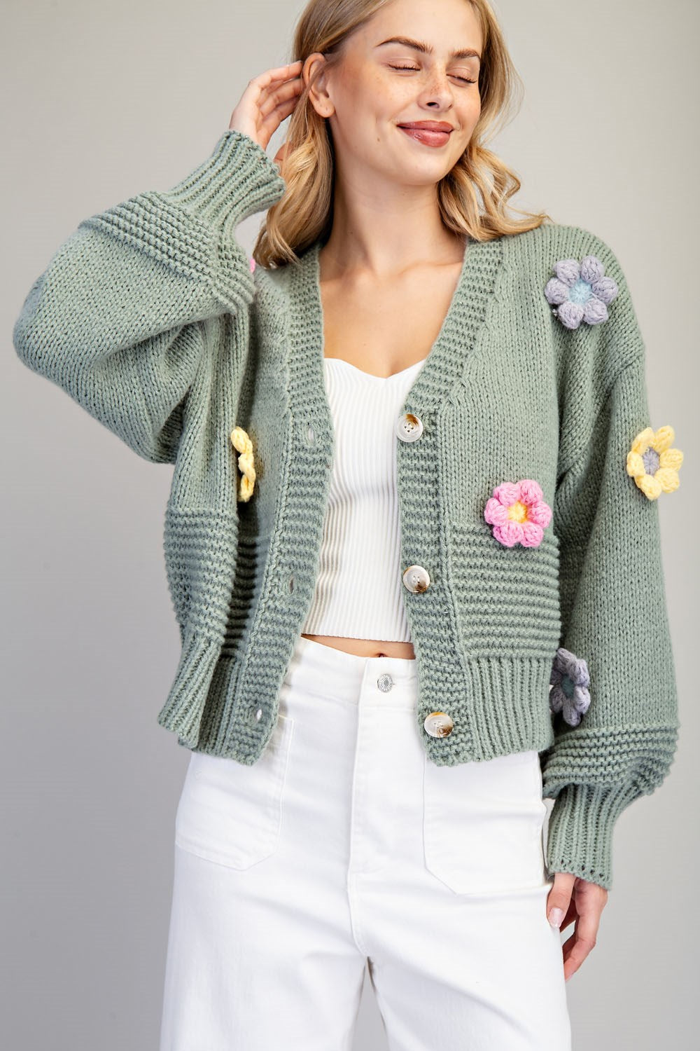 A woman wearing a sage green chunky knit cardigan with colorful crocheted flowers on the front and sleeves, styled over a white ribbed tank top and white pants against a solid gray studio background.