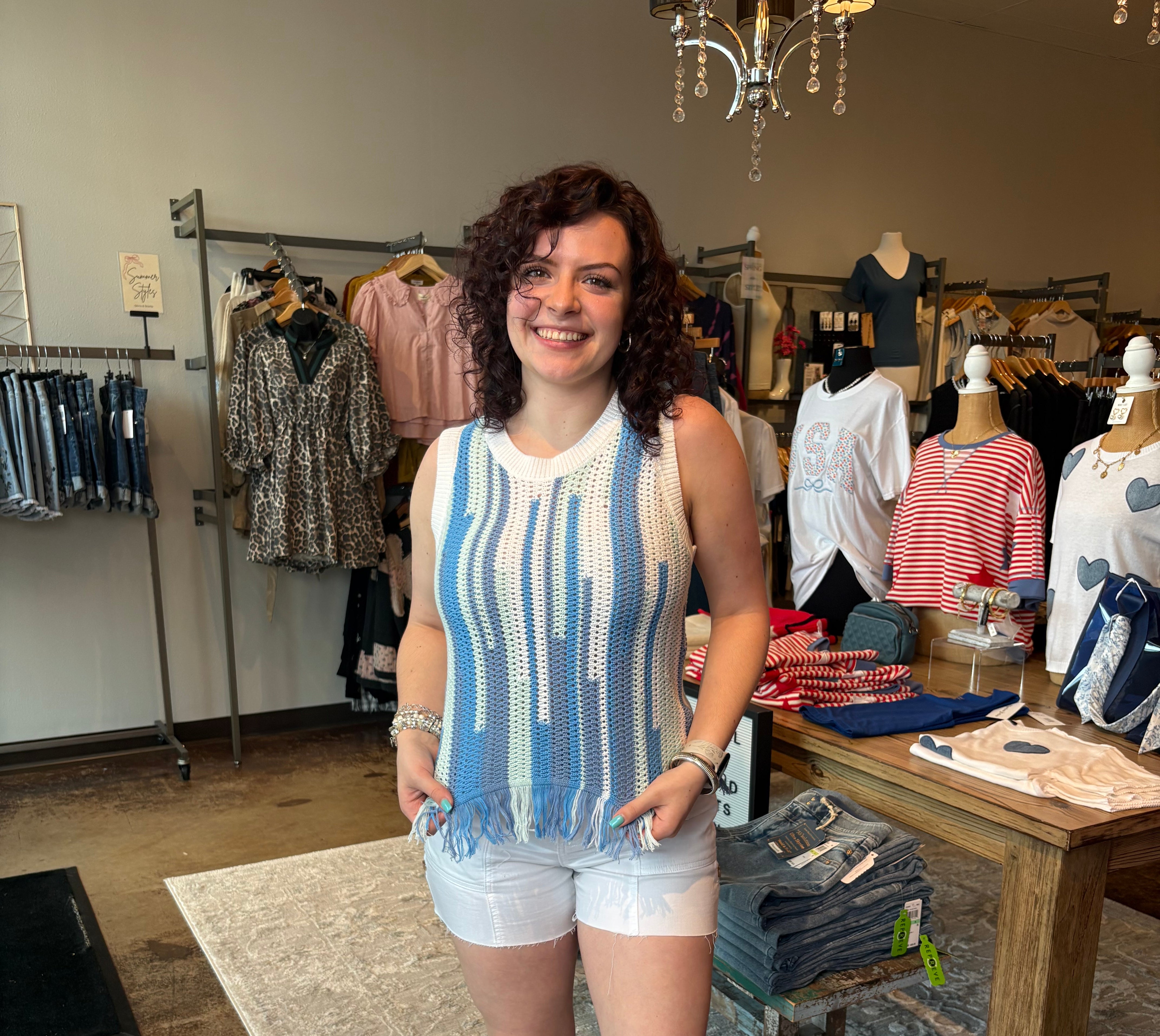 Woman standing in a clothing store wearing a blue and white striped sleeveless top and white shorts.