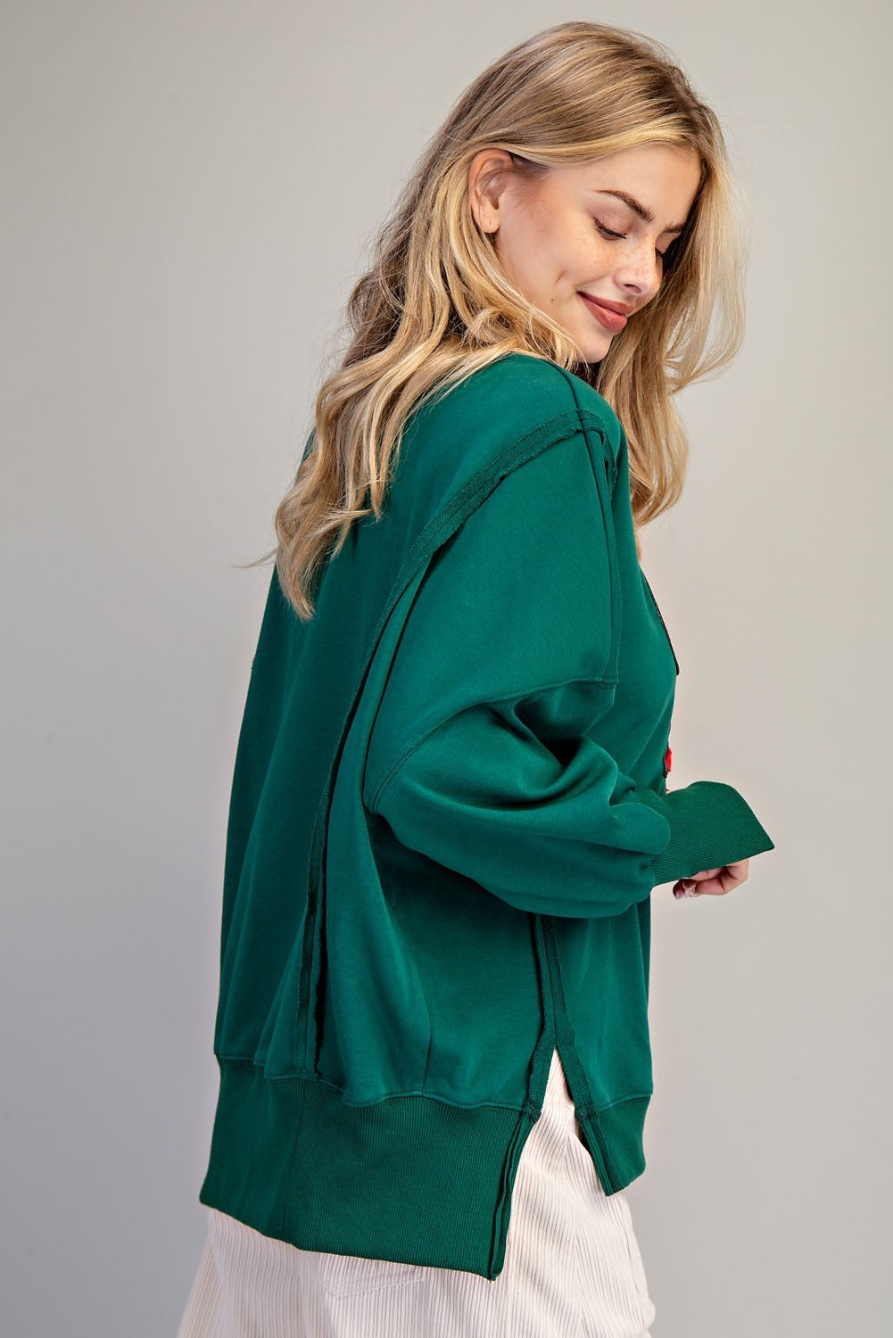 A woman wearing an oversized dark green long-sleeve top with side slits, paired with white pleated bottoms, photographed against a plain light gray studio background.