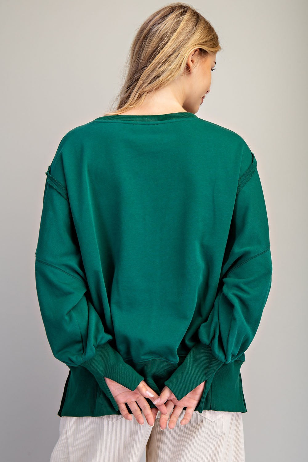 A woman viewed from behind wearing a dark green long-sleeve top with balloon sleeves and white pants, standing against a neutral gray background.