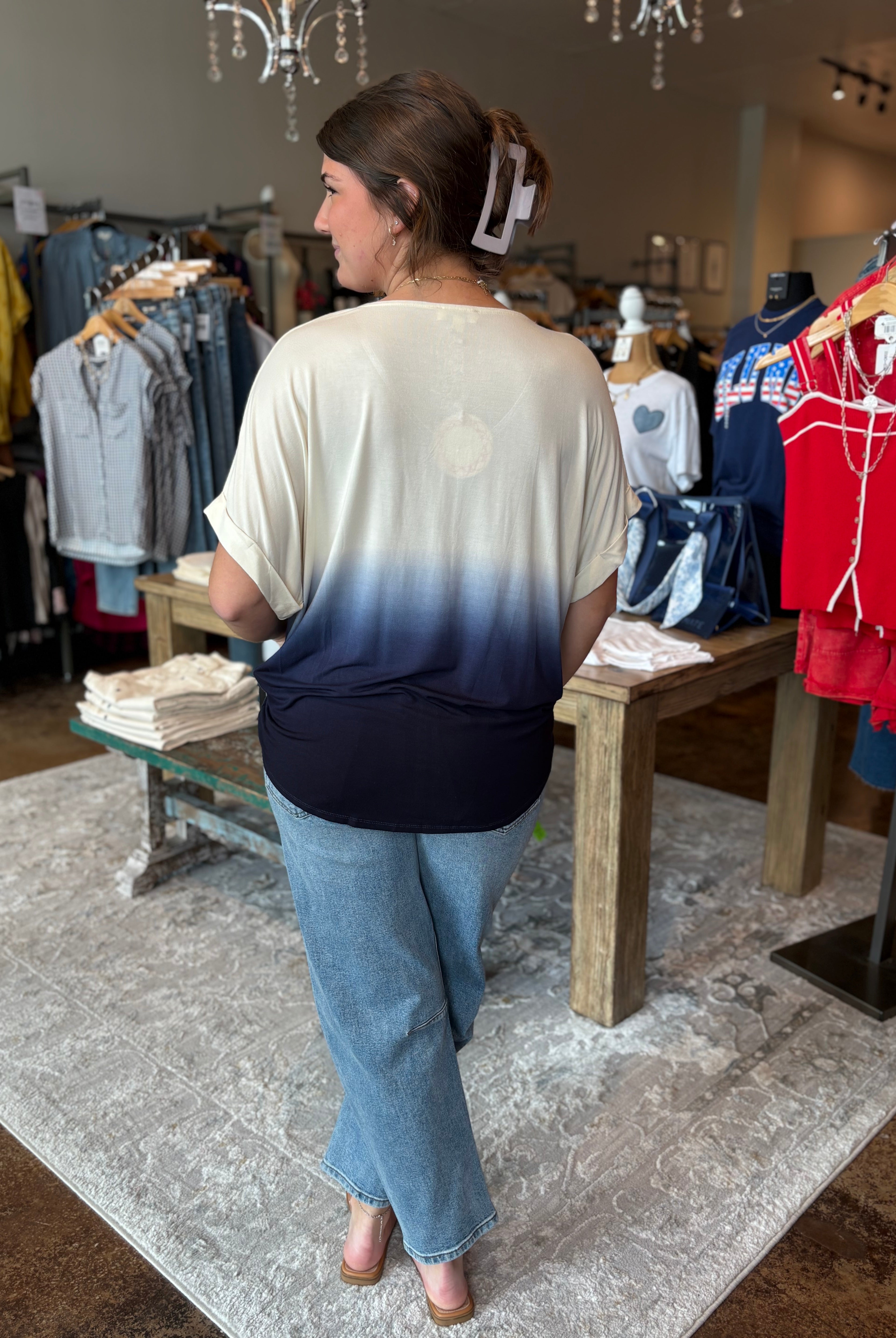 Woman in a store wearing an ombre shirt and jeans, looking at clothing on racks.