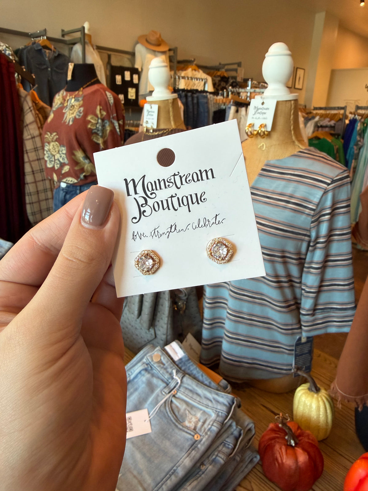 A hand holds a white display card featuring a pair of gold-toned stud earrings with clear stones. The card reads 'Mainstream Boutique' and 'Mr.