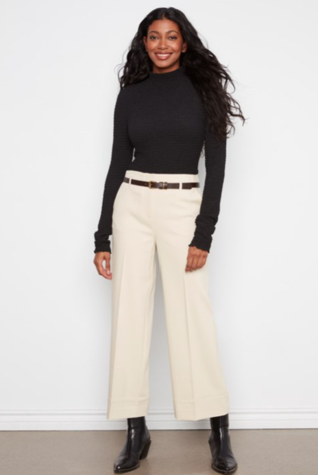 A woman modeling a black long-sleeve mock neck top and cream wide-leg cropped pants with a thin brown belt, paired with black ankle boots, against a white wall.