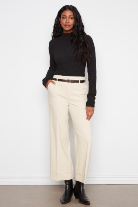 A woman modeling a black long-sleeve mock neck top and cream wide-leg cropped trousers with a thin brown belt, paired with black ankle boots, against a white studio background.