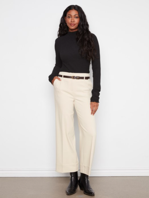 A woman modeling a black long-sleeve mock neck top and cream wide-leg cropped trousers with a thin brown belt, paired with black ankle boots, against a white studio background.