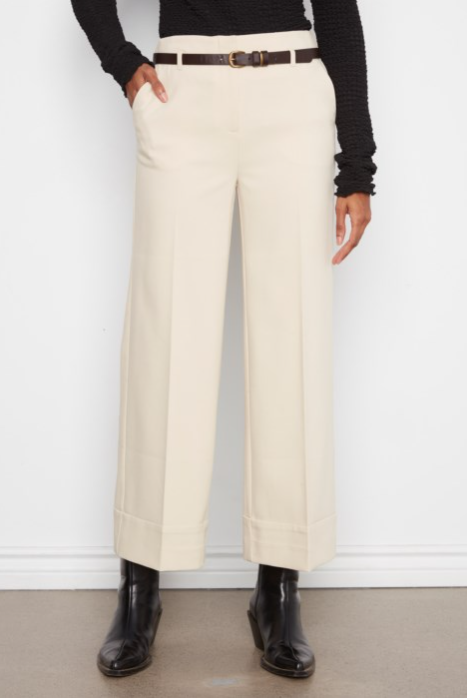 A person wearing high-waisted, wide-leg cropped trousers in a warm off-white/cream color, styled with a dark brown belt, a black ribbed long-sleeve top, and black leather ankle boots against a white wall.
