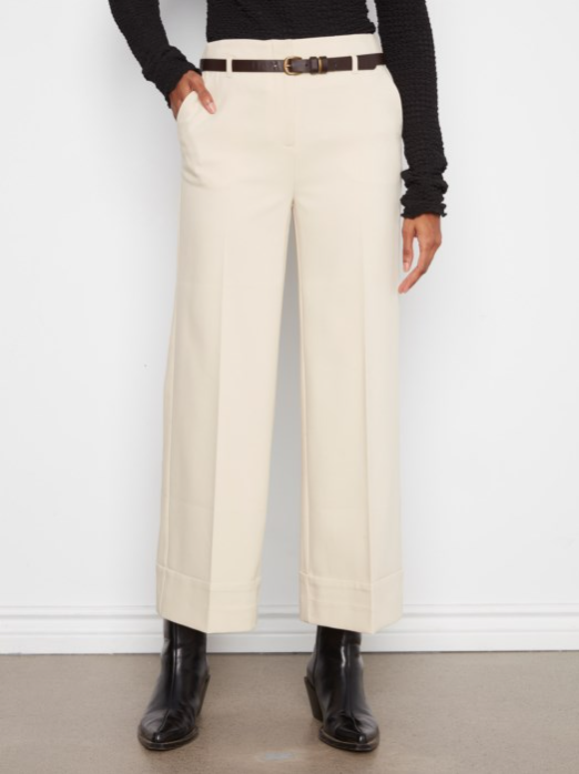 A person wearing high-waisted, wide-leg cropped trousers in a warm off-white/cream color, styled with a dark brown belt, a black ribbed long-sleeve top, and black leather ankle boots against a white wall.