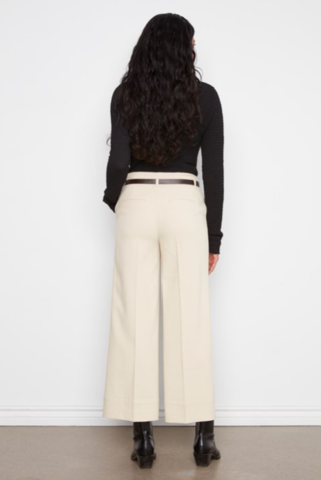 A woman shown from the back wearing a black long-sleeve top, cream wide-leg cropped trousers with a thin brown belt, and black ankle boots, standing against a white wall.