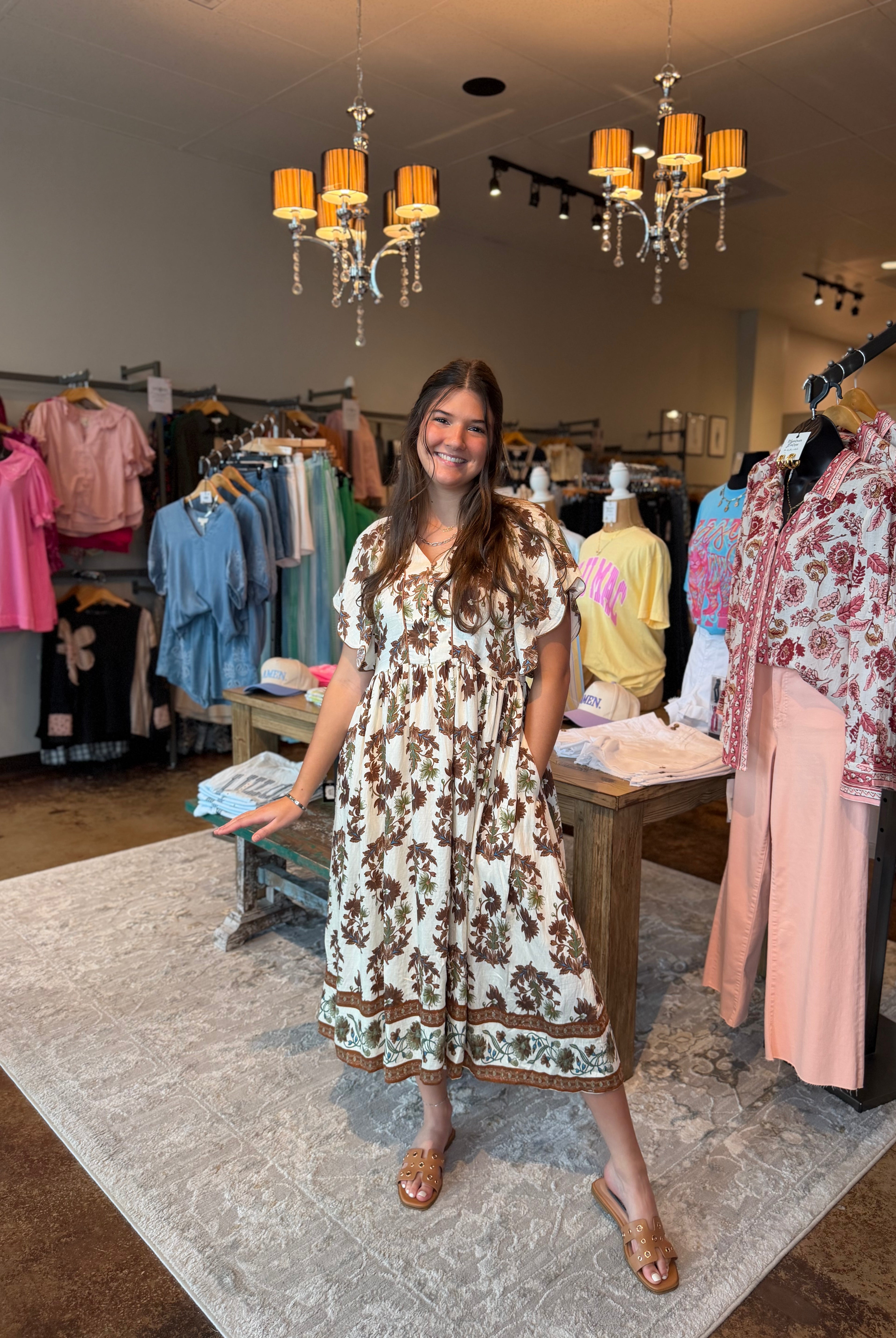 A woman modeling a brown and cream floral print midi dress inside a boutique clothing store, surrounded by racks of colorful apparel and hanging chandeliers.