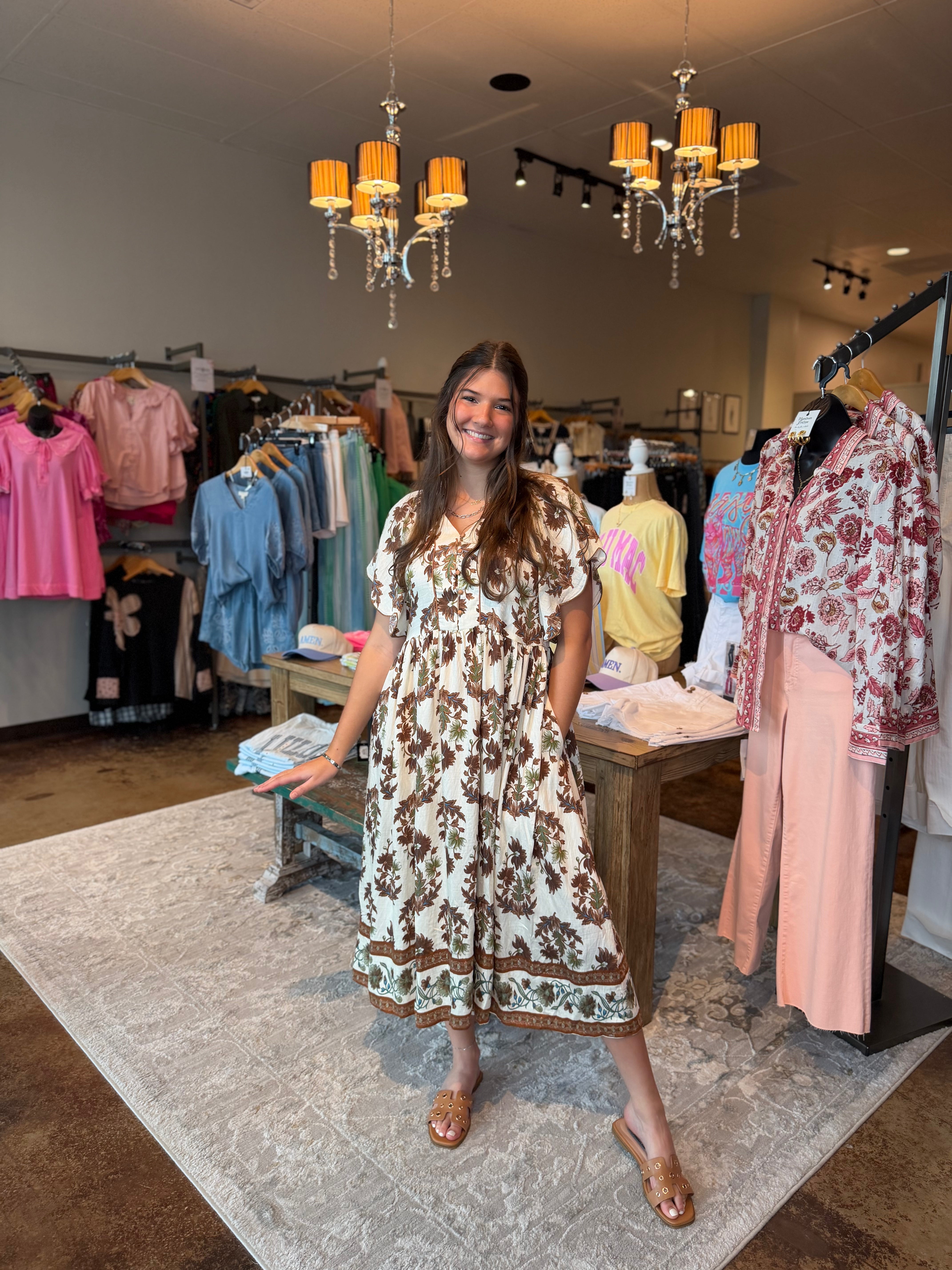 A woman modeling a brown and cream floral print midi dress inside a boutique clothing store, surrounded by racks of colorful apparel and hanging chandeliers.