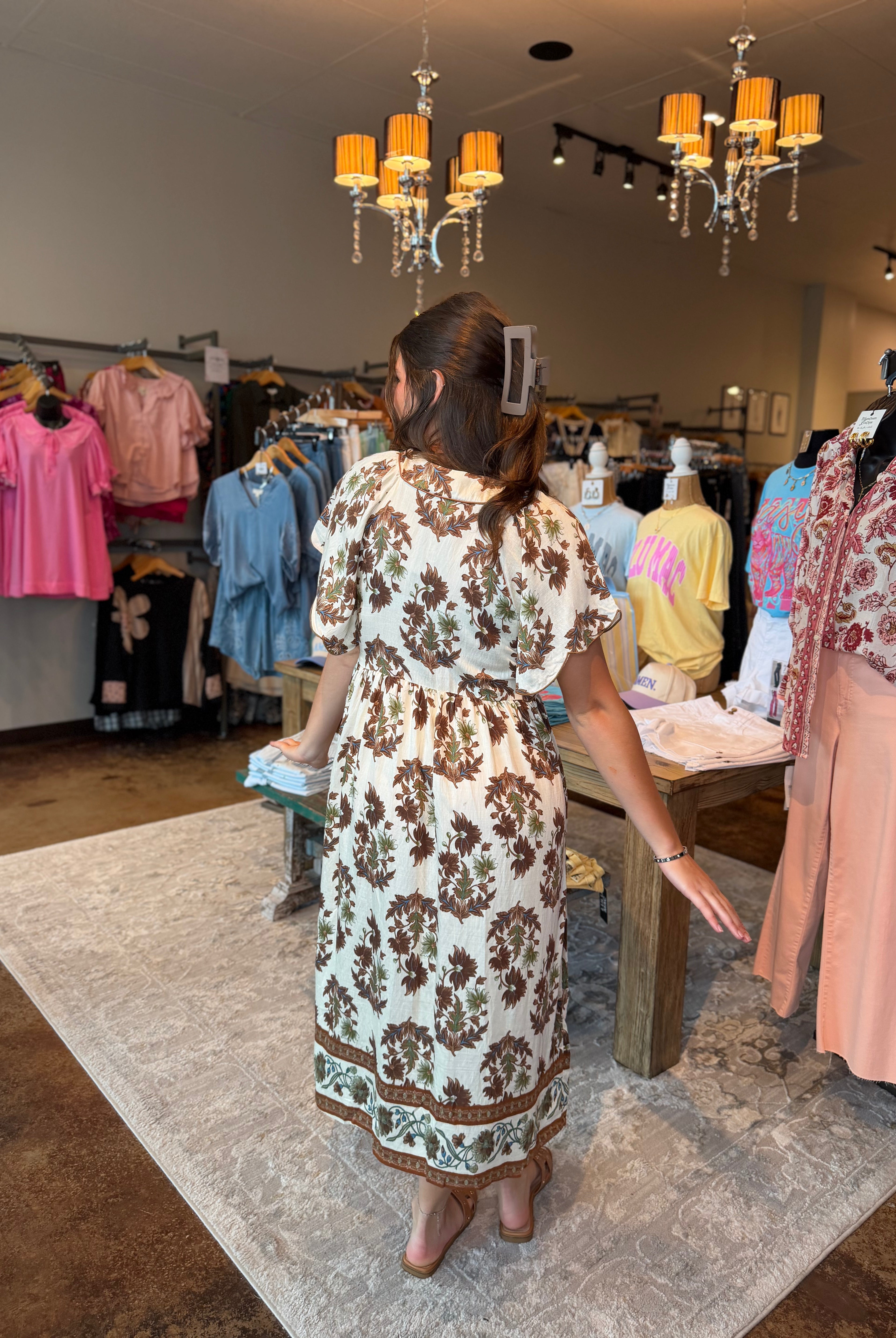 A woman viewed from behind wearing a cream-colored dress with a brown floral pattern, standing inside a clothing boutique on a patterned rug.