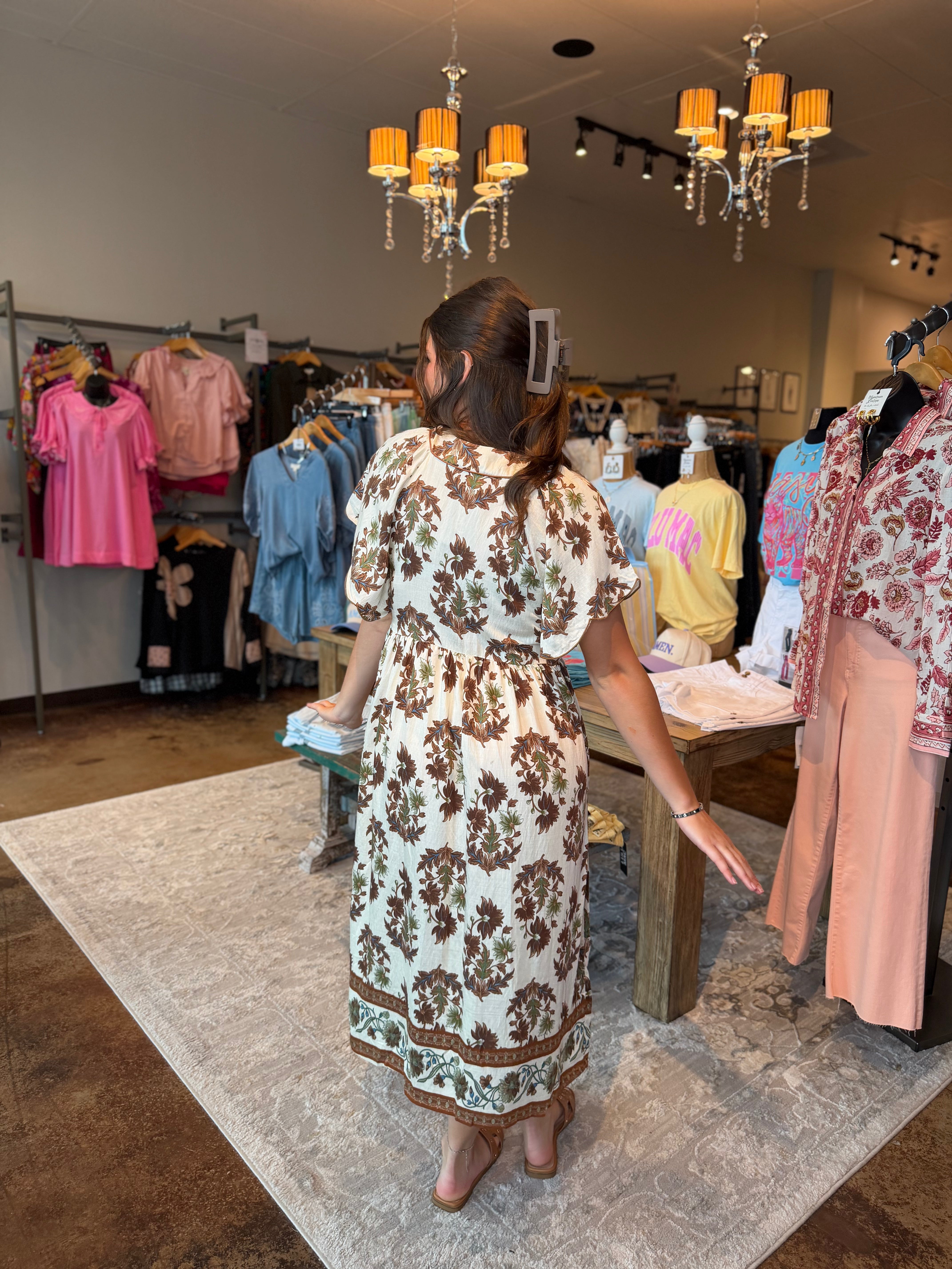 A woman viewed from behind wearing a cream-colored dress with a brown floral pattern, standing inside a clothing boutique on a patterned rug.