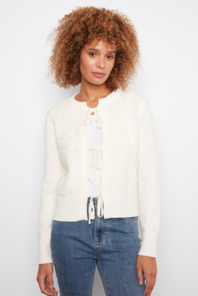 A woman with curly brown hair wearing a cream-colored knit cardigan with lace-up front details and blue jeans, standing against a plain white studio background.