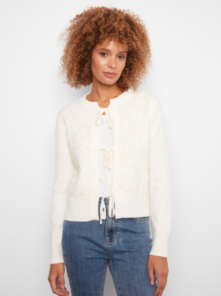 A woman with curly brown hair wearing a cream-colored knit cardigan with lace-up front details and blue jeans, standing against a plain white studio background.
