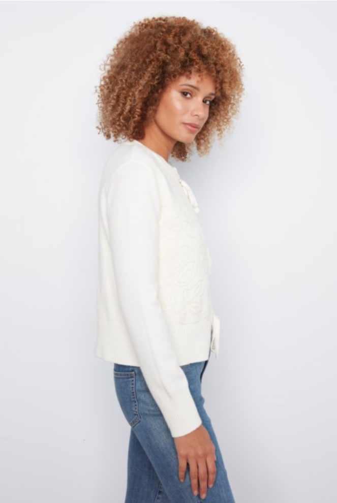 A woman with curly light brown hair is shown in profile wearing a white knit cardigan with lace-up detailing and blue jeans, set against a plain white studio background.