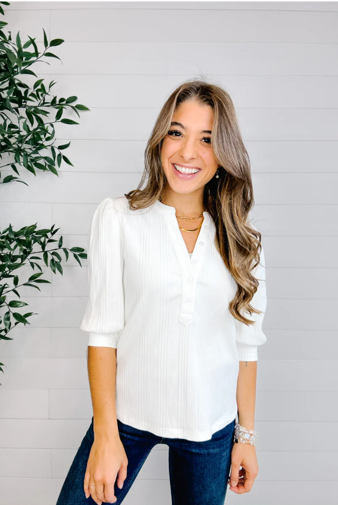 Woman wearing a white blouse and blue jeans standing next to a plant against a light gray wall.
