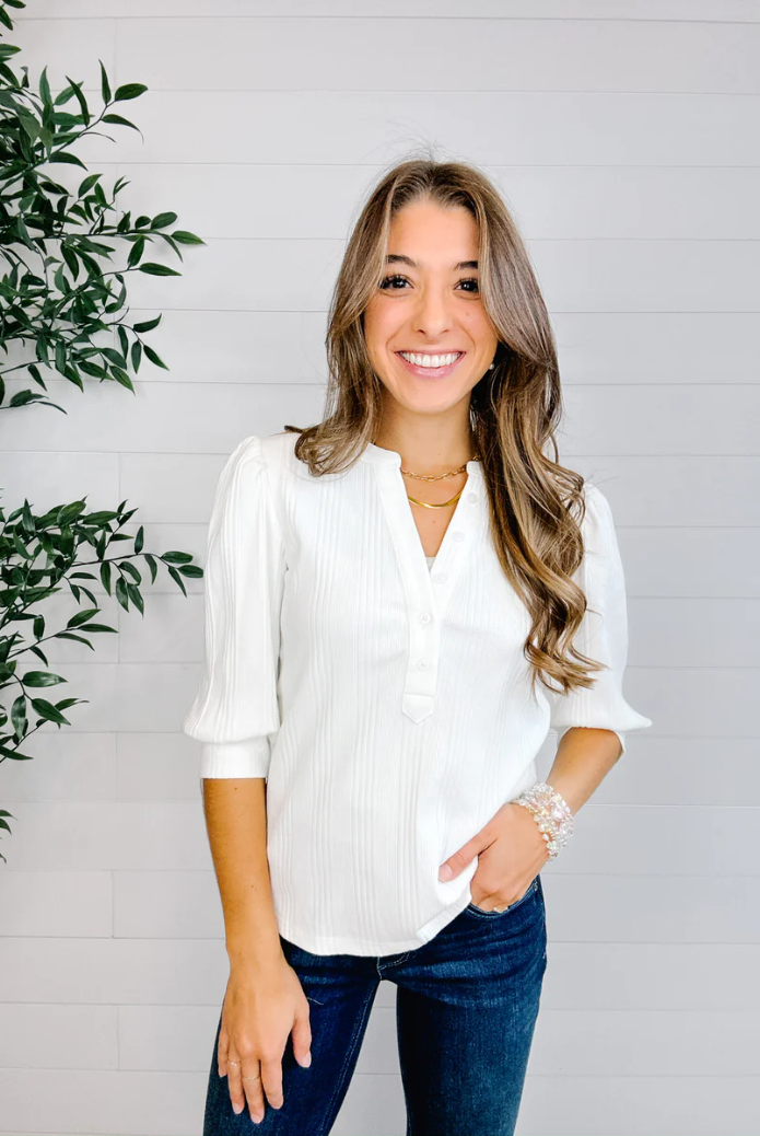 Woman wearing a white blouse and blue jeans standing against a light gray wall with a plant to the left.