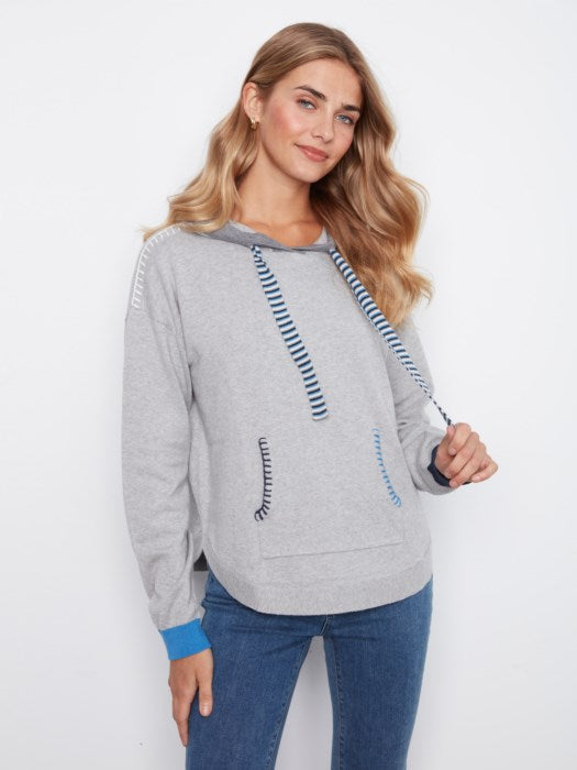 A woman wearing a light gray heathered hoodie with blue and white striped drawstrings, blue striped pocket trim, and a bright blue ribbed cuff, paired with blue jeans, against a plain white background.