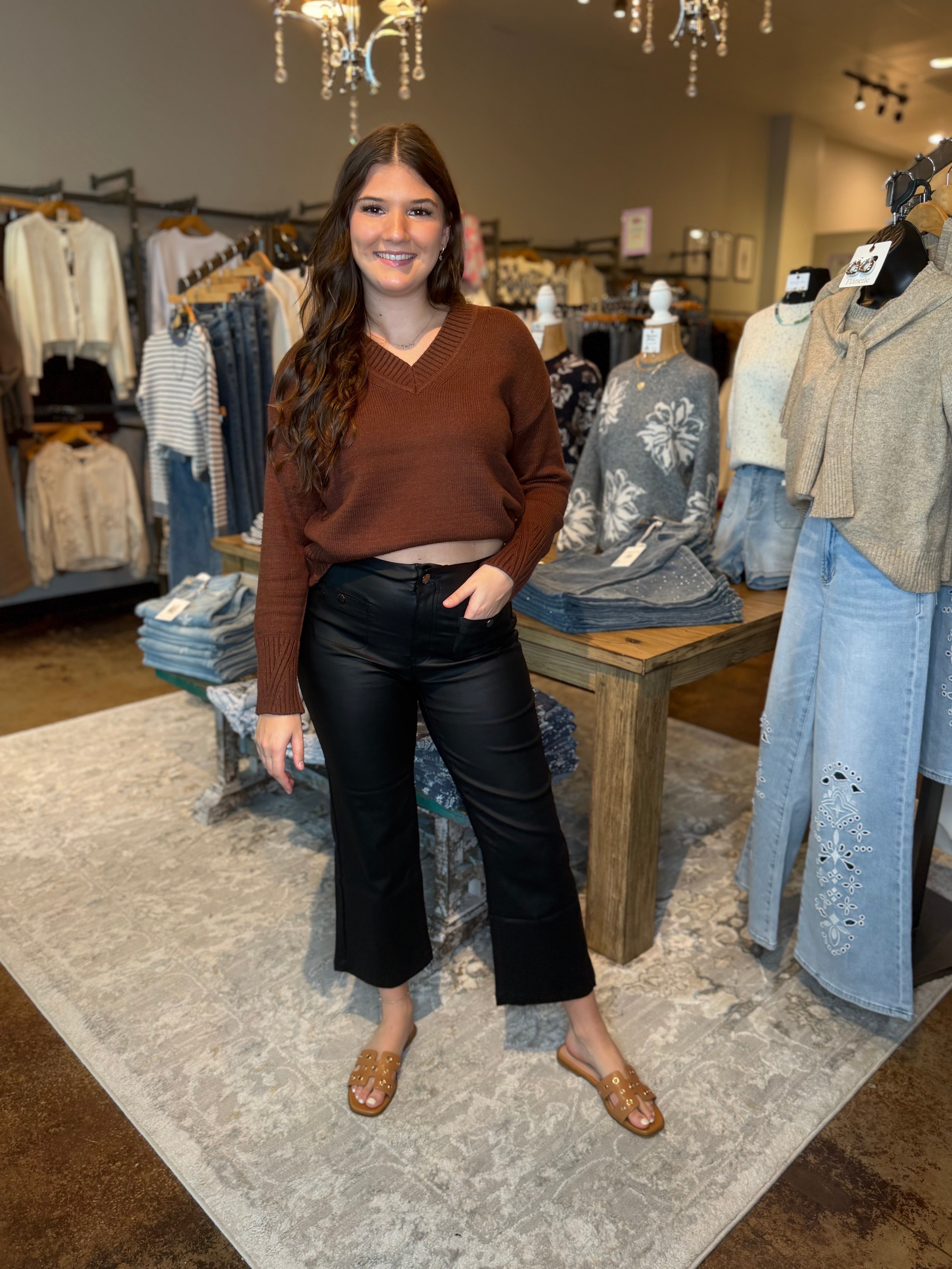 A woman stands in a clothing boutique wearing a brown long-sleeve cropped V-neck sweater, black cropped pants, and brown mule sandals. The boutique has mannequins with sweaters, clothing racks, and a patterned rug.