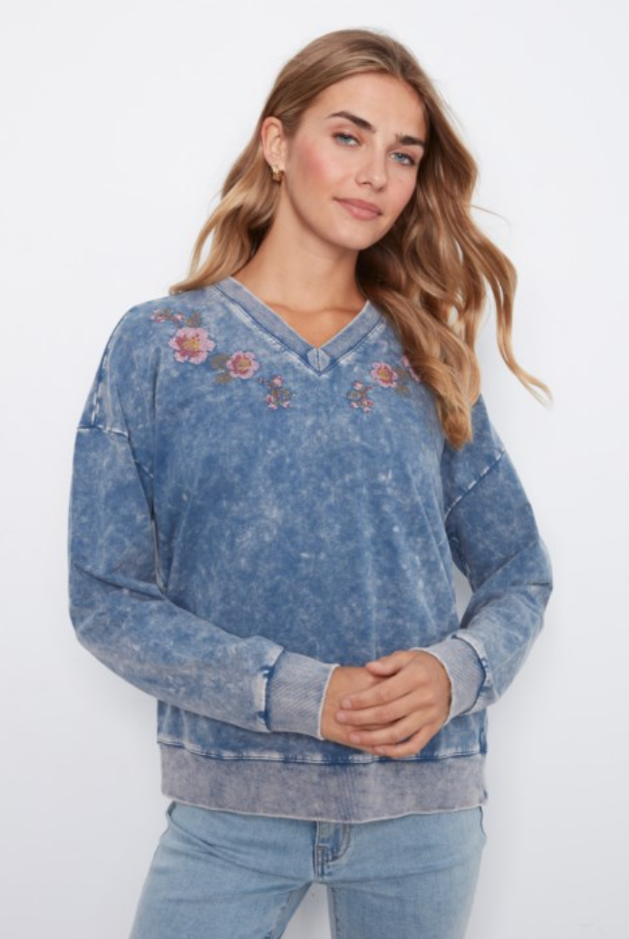 A woman wearing a blue acid-wash V-neck sweatshirt with floral embroidery on the chest, paired with light-wash jeans, against a plain white background.