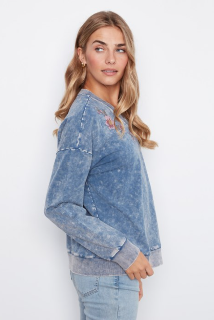 A woman modeling a blue acid-wash long-sleeve sweatshirt with a small embroidered graphic on the chest, paired with light-wash jeans against a plain white background.