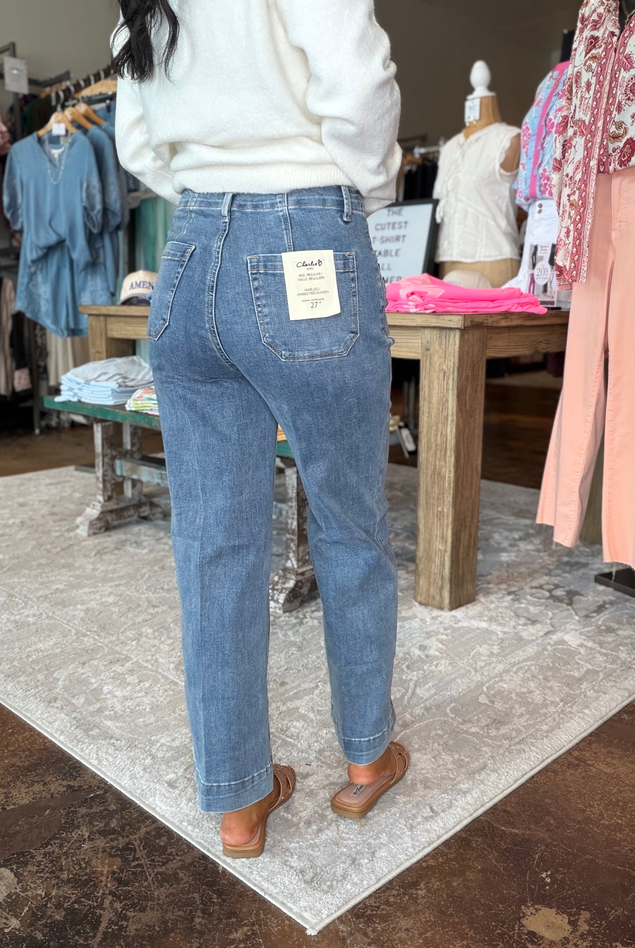 A person standing in a clothing store wearing light-wash blue jeans with a white sweater and brown flat shoes, shown from the back. The jeans have a visible back pocket label.