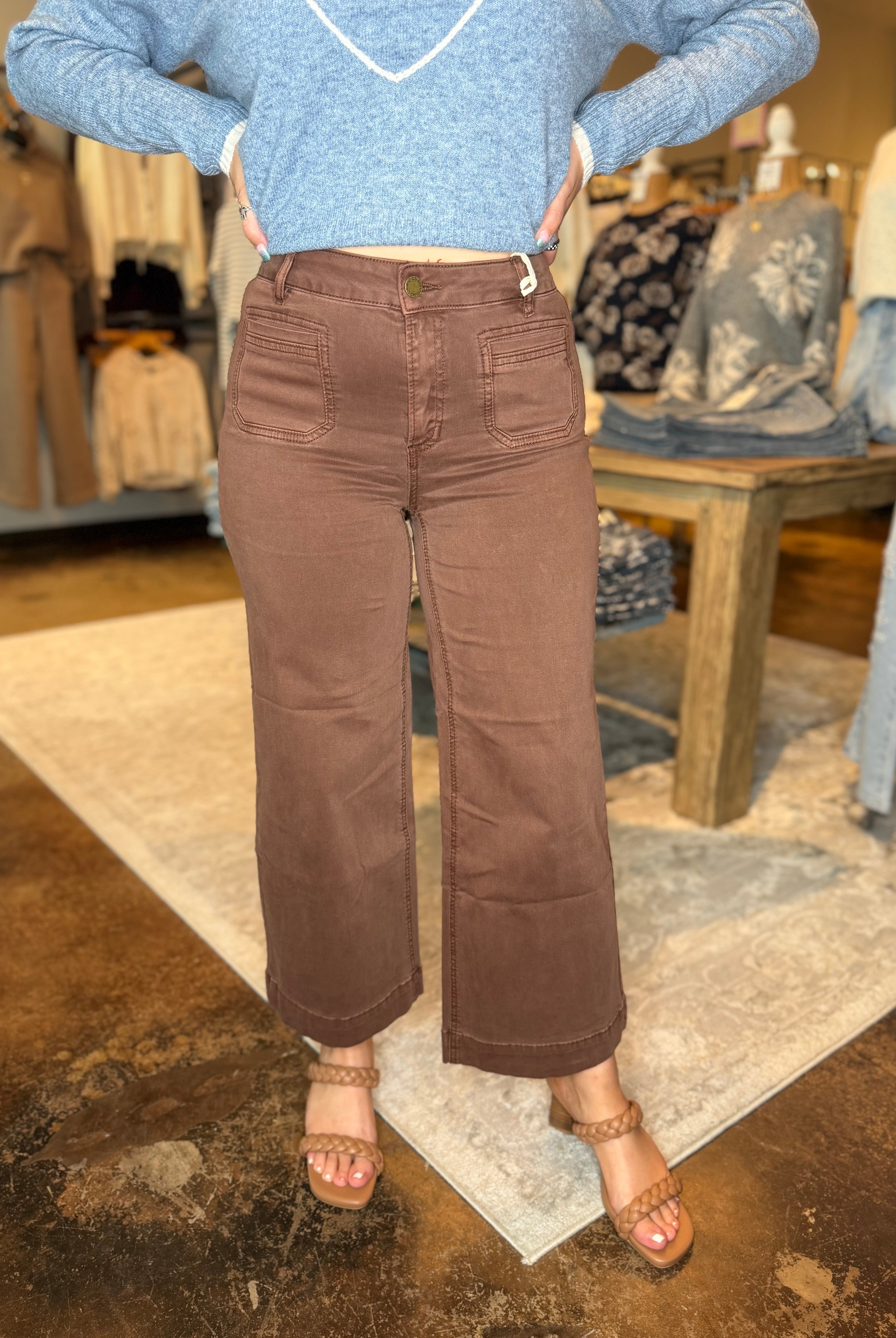 A person wearing brown wide-leg cropped pants with a light blue knit sweater and tan heeled sandals, standing in a clothing store.