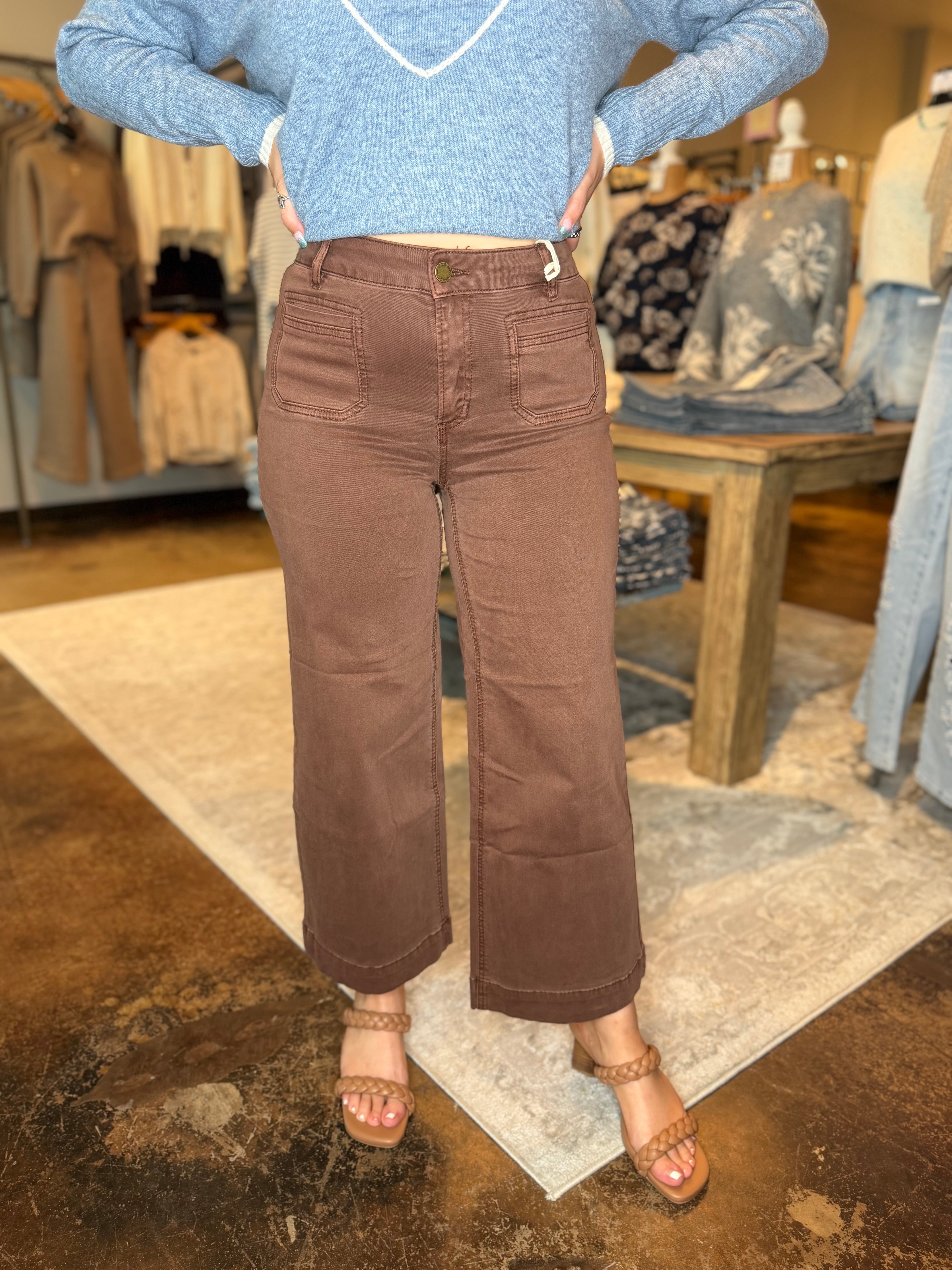 A person wearing brown wide-leg cropped pants with a light blue knit sweater and tan heeled sandals, standing in a clothing store.