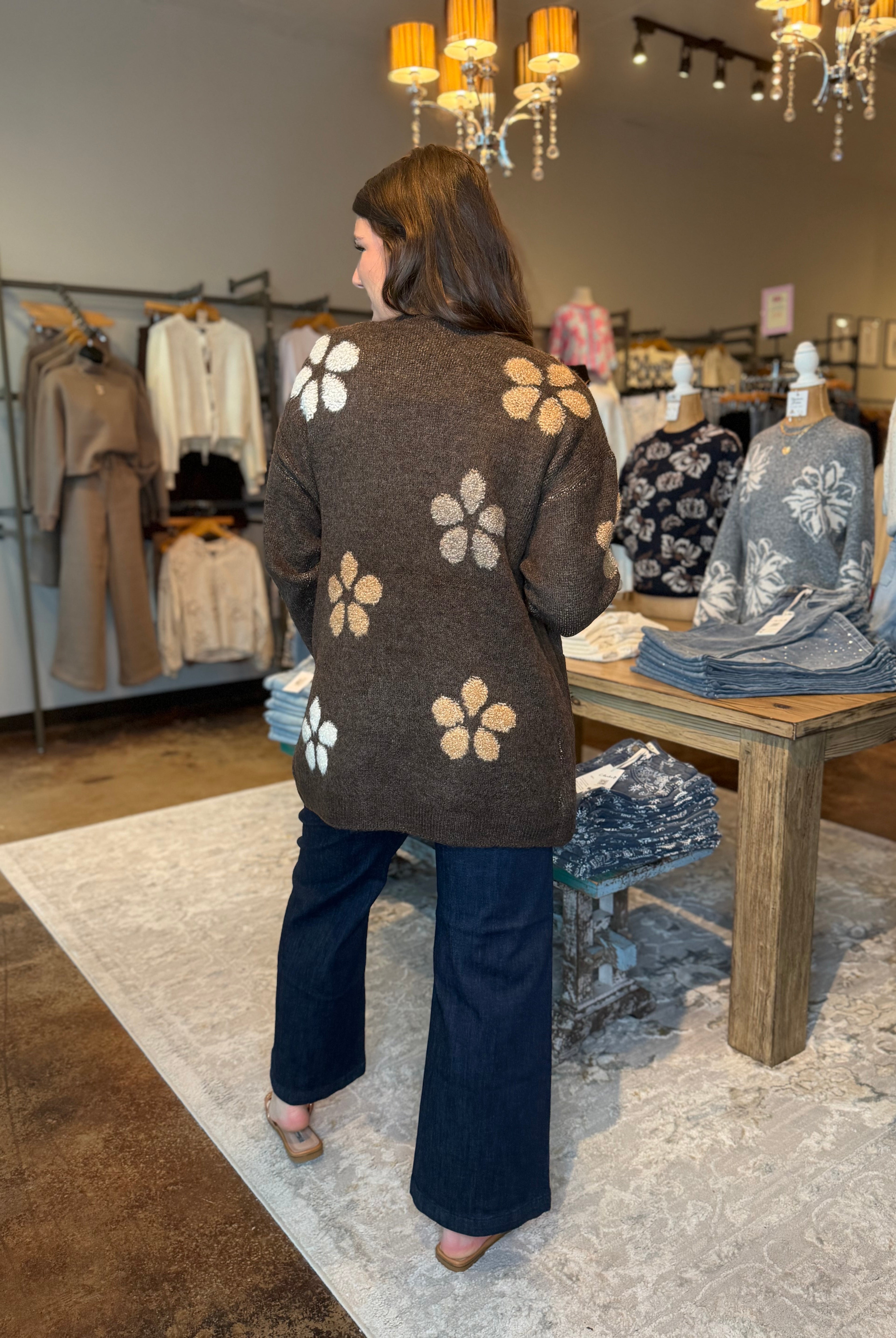 A woman viewed from behind wearing a brown knit cardigan with large white and tan floral motifs, paired with dark wide-leg pants in a boutique clothing store.