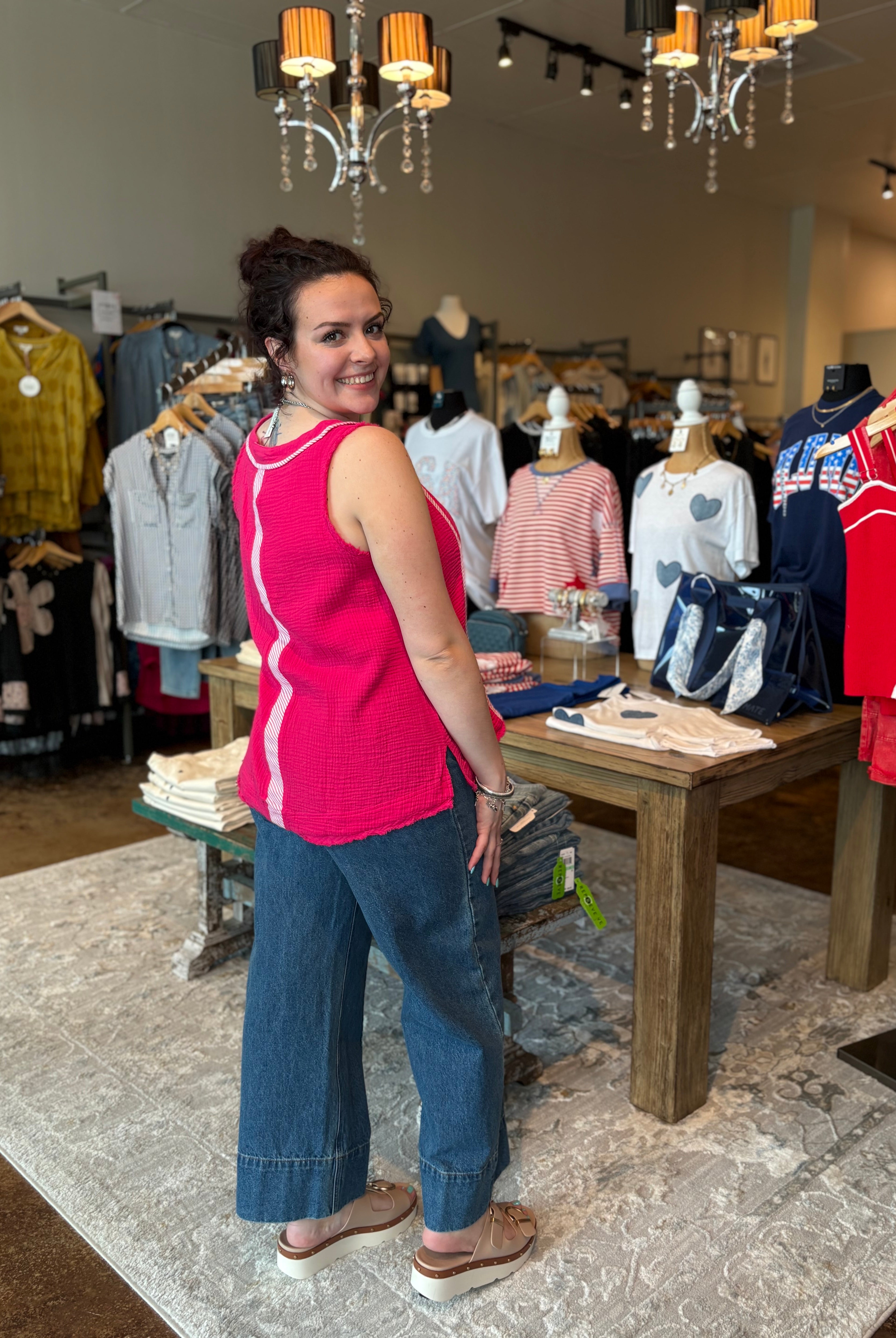 Woman in a pink top and blue jeans standing in a clothing store with racks of clothes and a table.