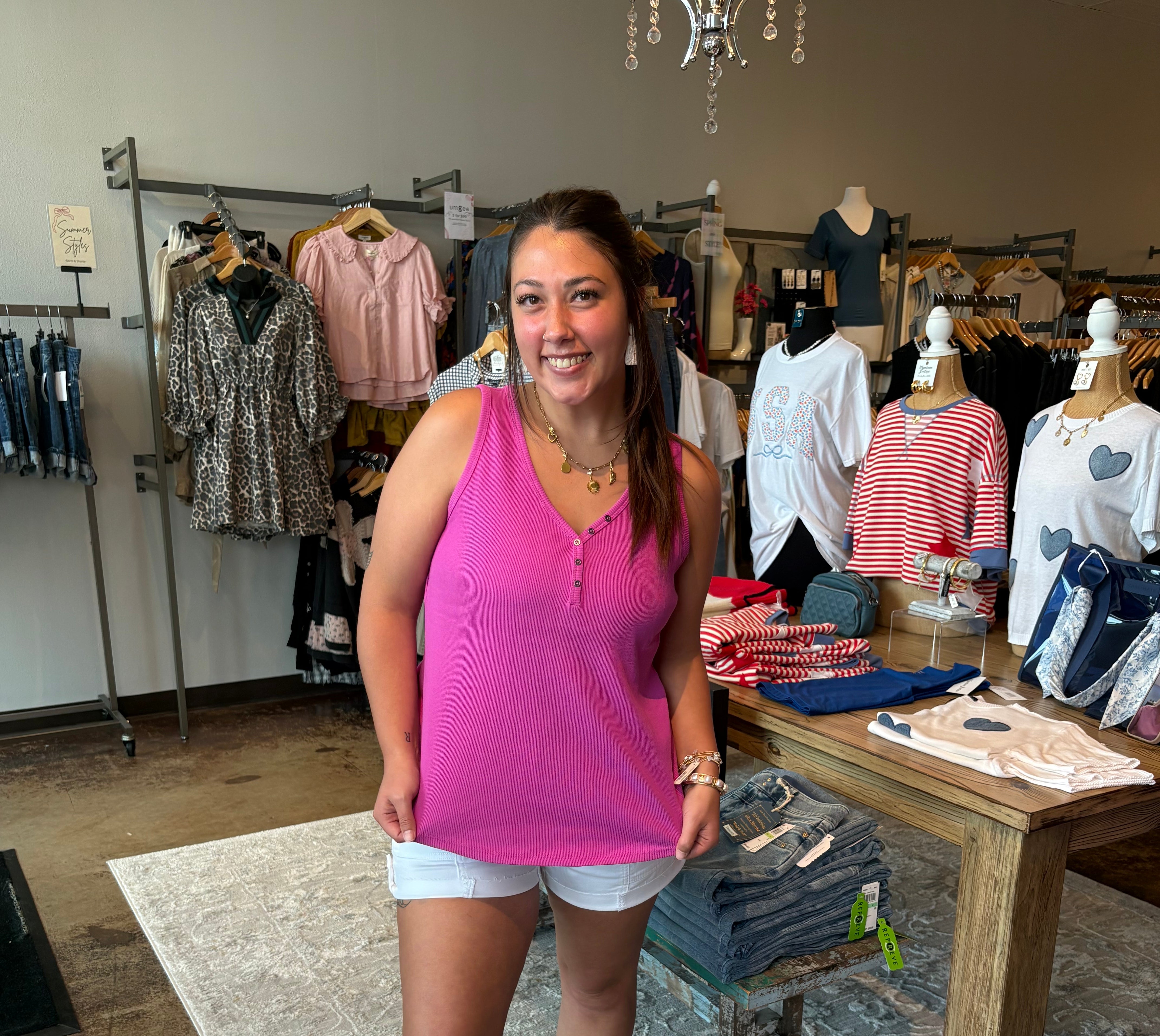 Woman in a pink tank top standing in a clothing store with racks of clothes in the background.