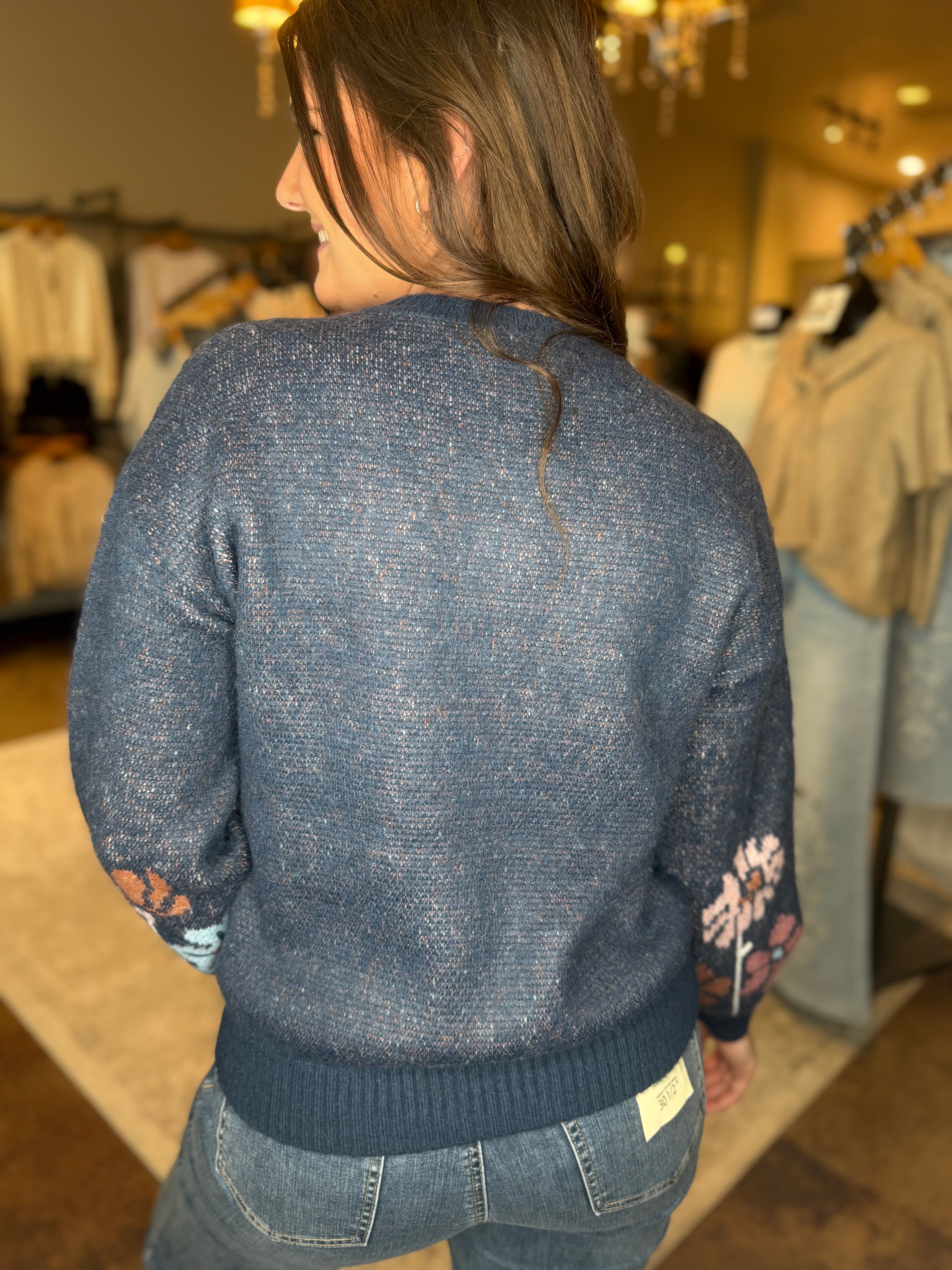 A woman viewed from behind wearing a textured blue knit sweater with floral embroidery on the sleeves, standing inside a clothing store.