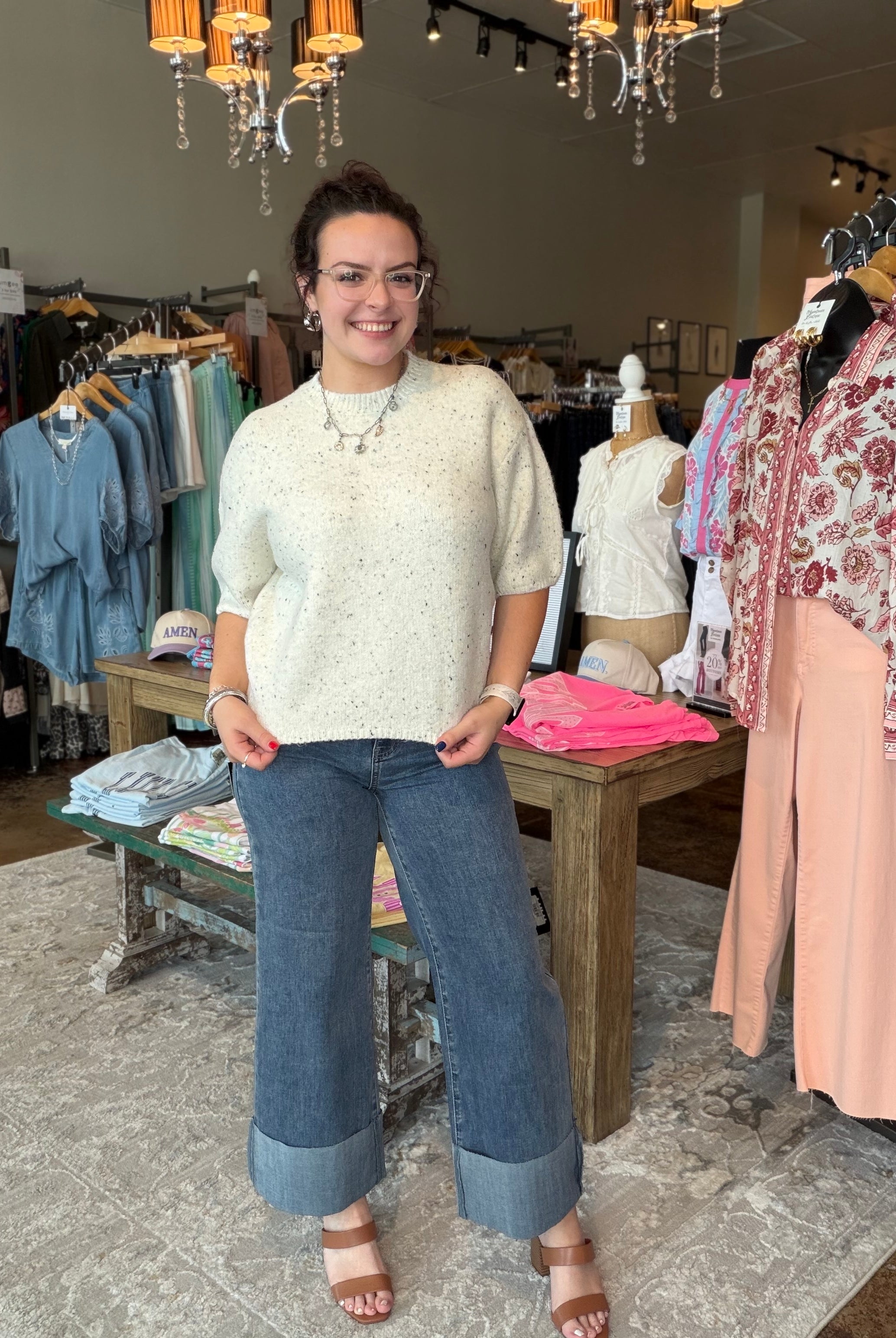 A woman stands in a clothing boutique wearing a cream-colored short-sleeve knit top with subtle speckles, blue wide-leg cuffed jeans, and brown heeled sandals. Clothing racks, mannequins, and store fixtures are visible in the background.