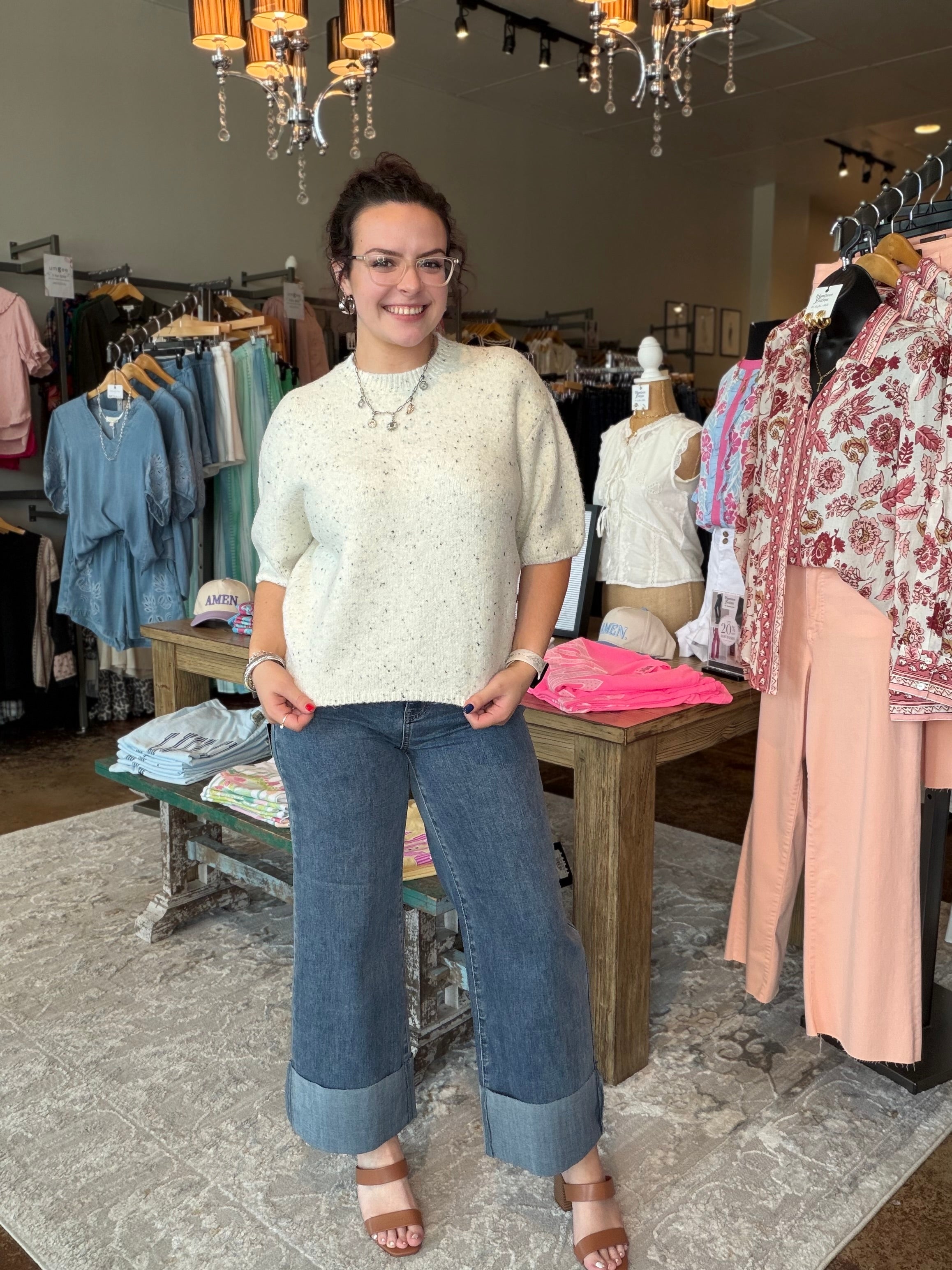 A woman stands in a clothing boutique wearing a cream-colored short-sleeve knit top with subtle speckles, blue wide-leg cuffed jeans, and brown heeled sandals. Clothing racks, mannequins, and store fixtures are visible in the background.