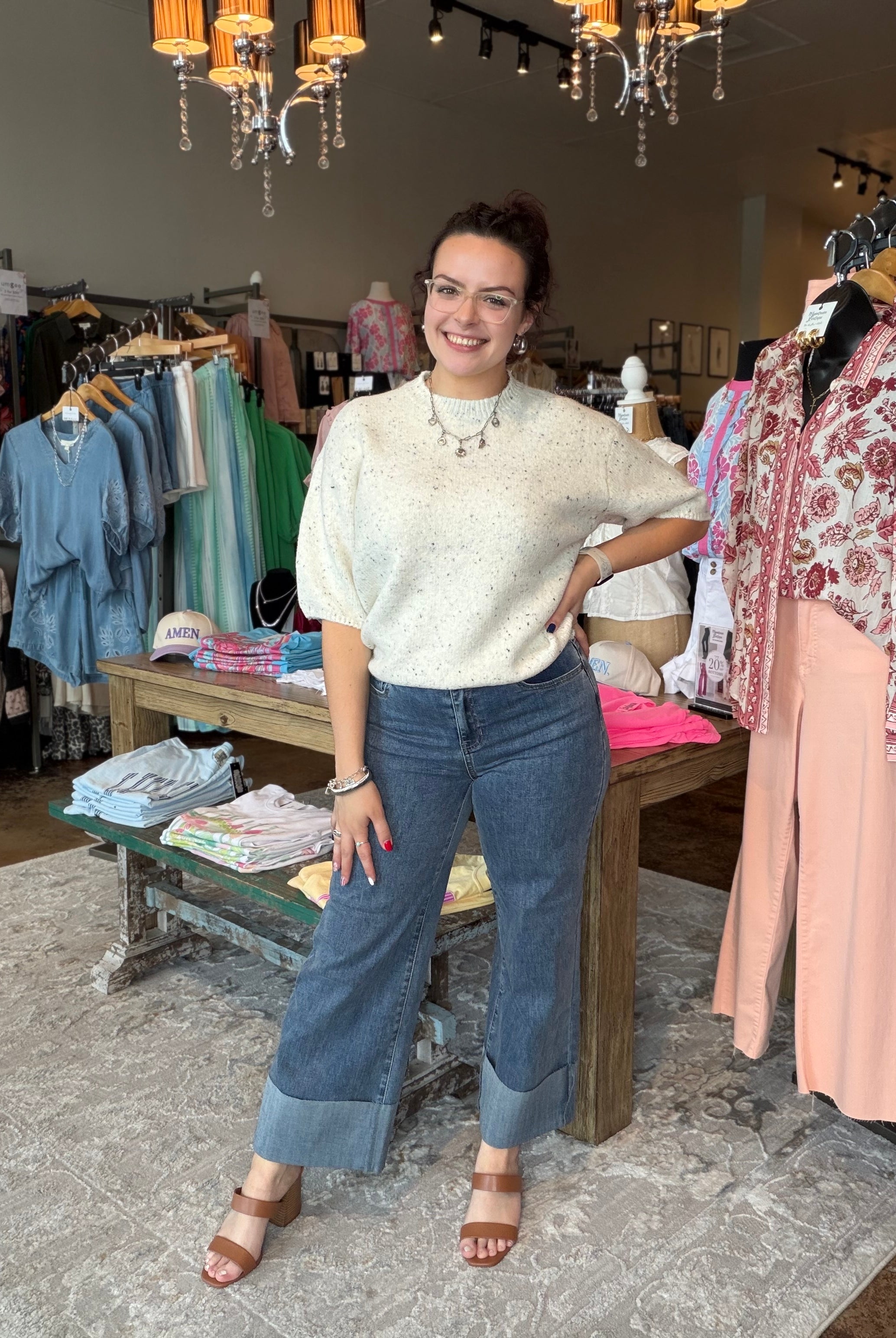 A woman posing in a boutique store wearing a cream-colored textured short-sleeve top and blue wide-leg cuffed jeans.