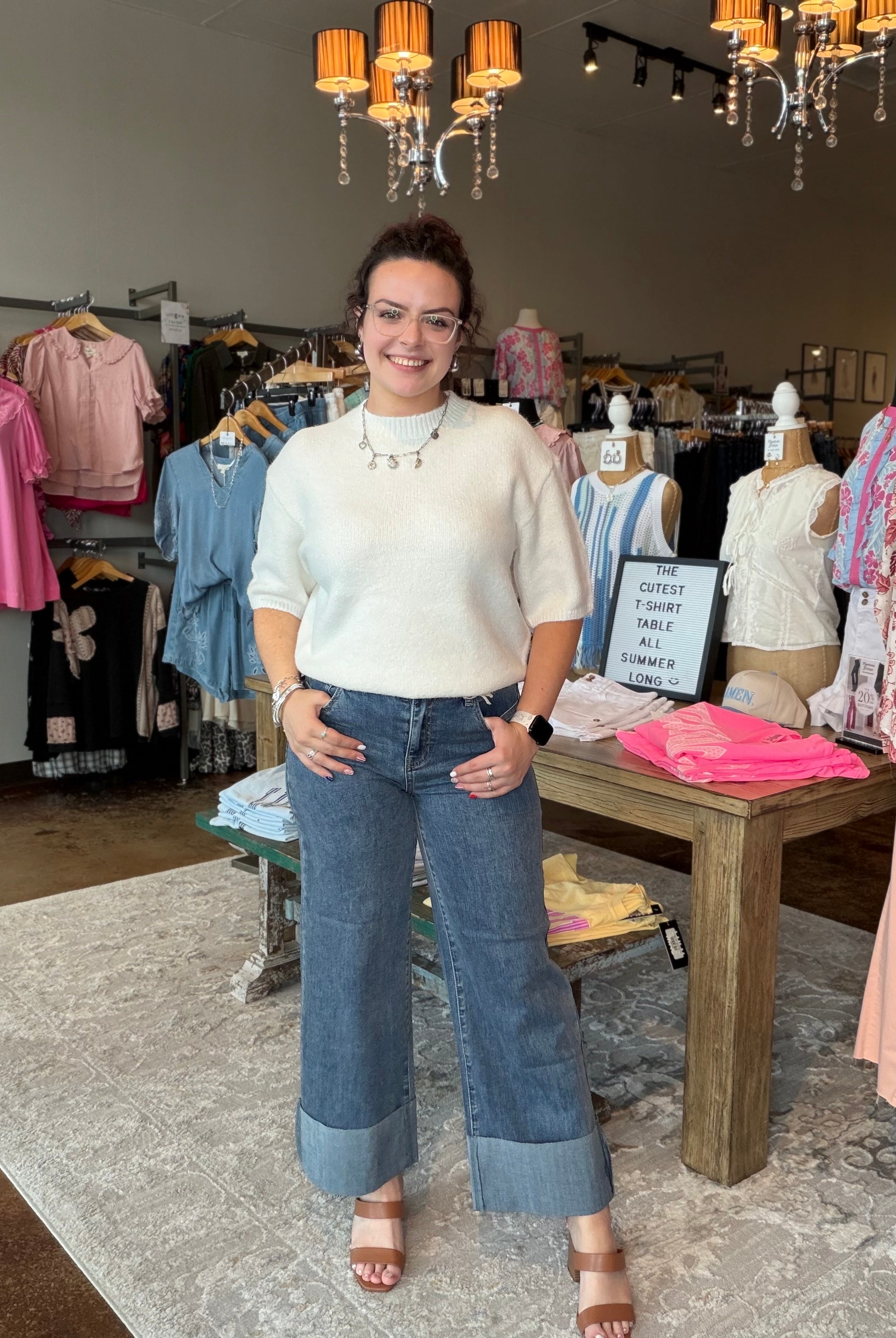 A woman stands inside a clothing boutique wearing a cream short-sleeve knit top and wide-leg blue jeans with rolled hems. The boutique has clothing racks, mannequins, a wooden display table, and hanging chandeliers in the background.